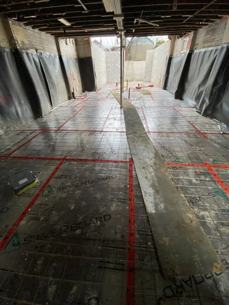 Construction site of a building interior with concrete floor and utility markings, partially enclosed with black plastic sheets on the walls, and an opening showing outdoor yard with trees and neighboring buildings.
