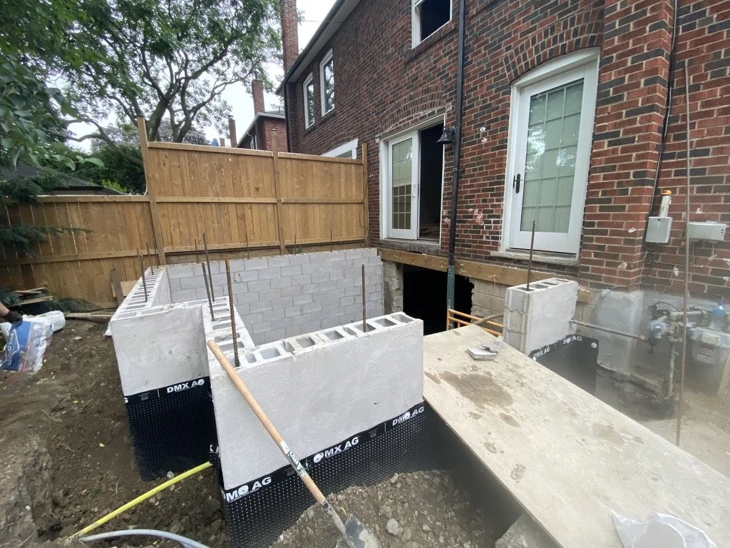 Construction site with new concrete foundation and cinder blocks for a home addition next to a brick house, with wooden fencing and trees in the background.