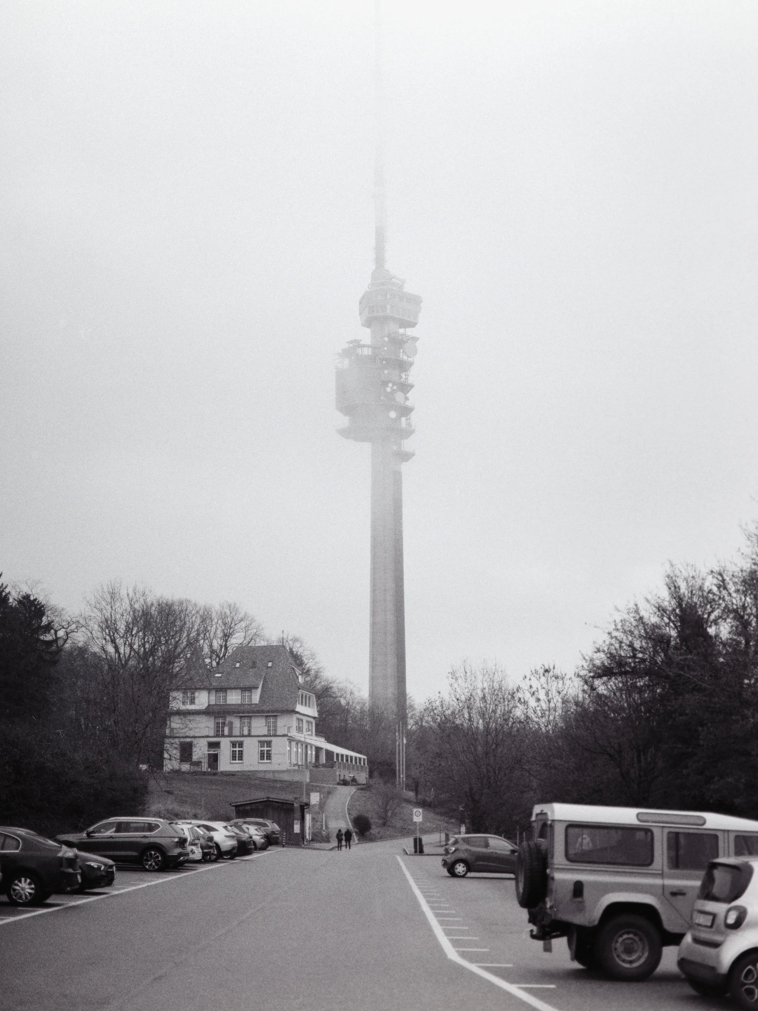 A foggy scene featuring a tall communication tower with multiple platforms above a residential building and cars parked in a lot, with trees around in St. Chrischona.