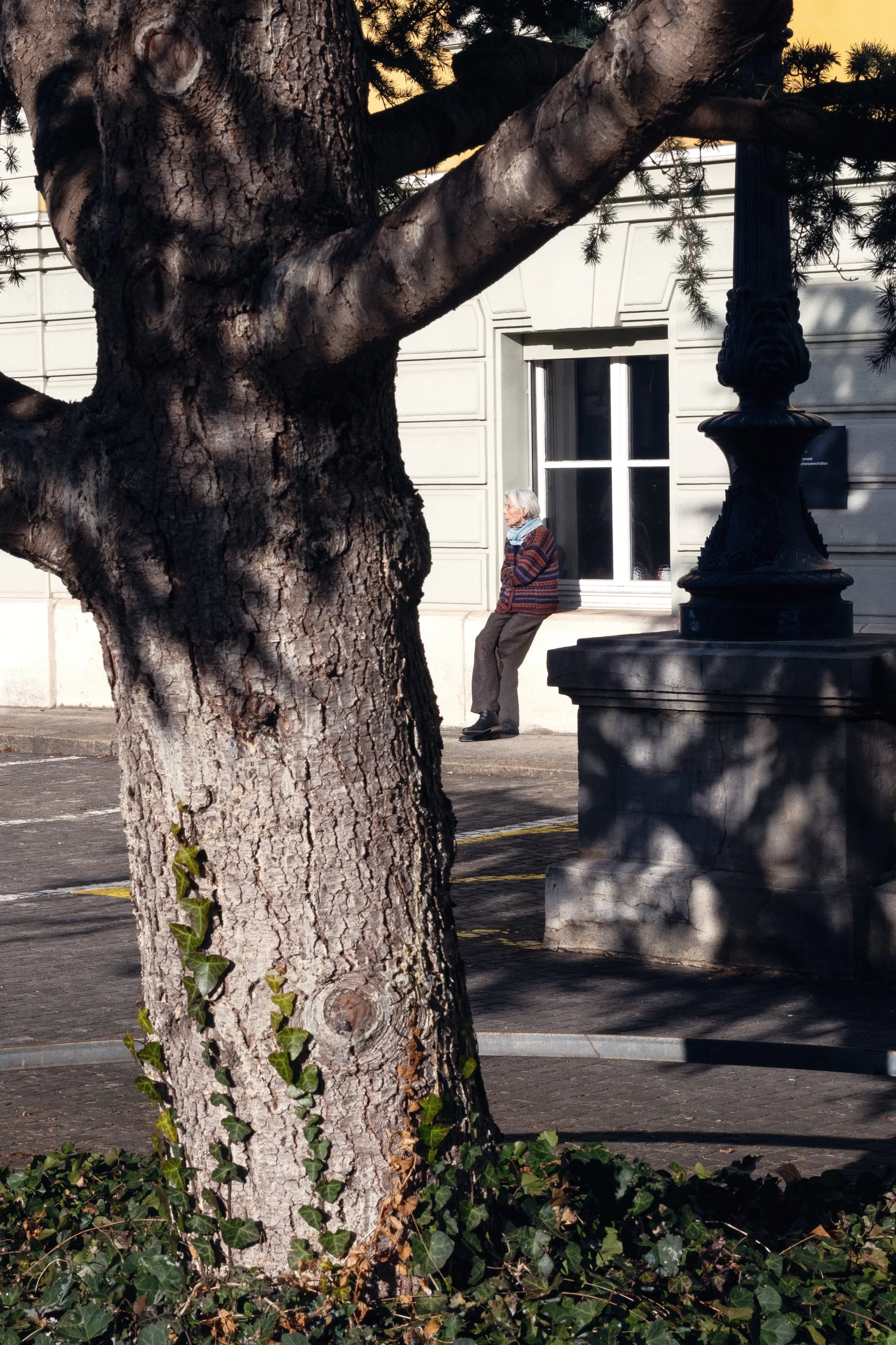 A woman sitting outside a building and smoking framed by a tree and a light post