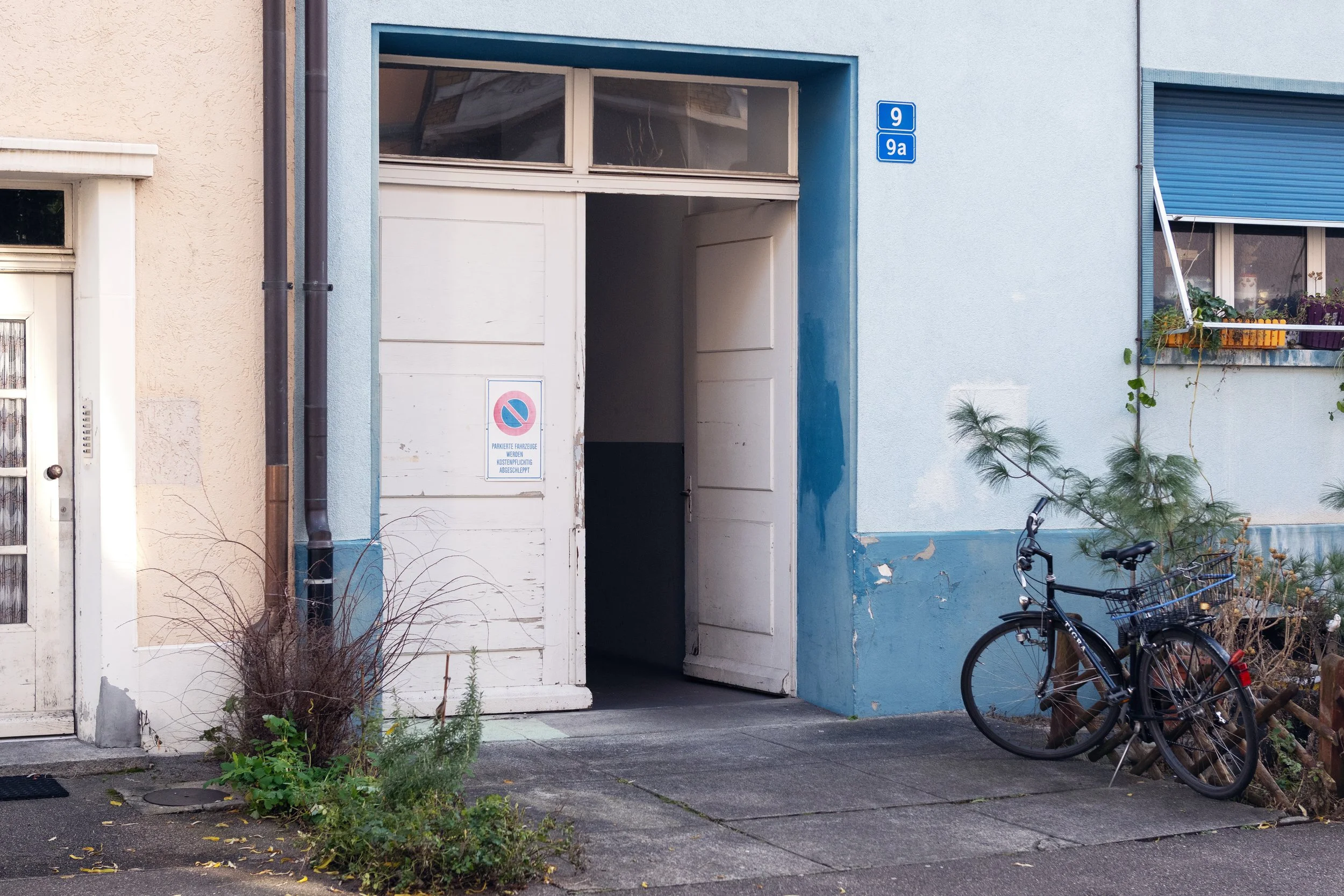 Building entrance with a blue and white color scheme, a bicycle parked against the wall, and some plants near the sidewalk in Basel, Switzerland.