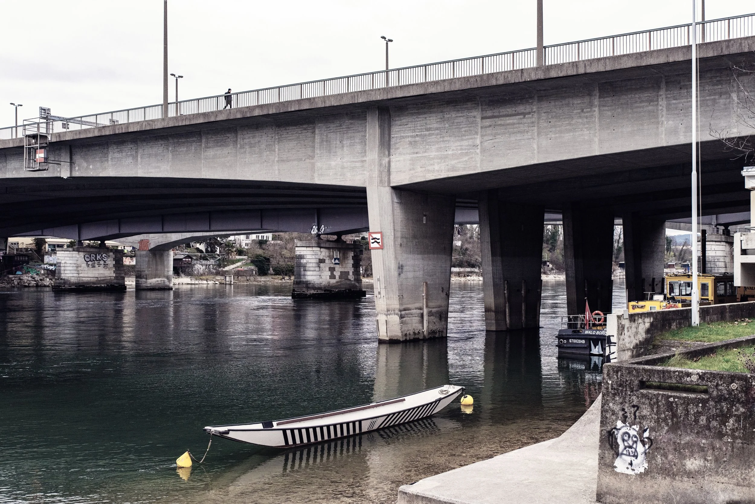 A concrete bridge over the Rhine in Basel with a small boat moored in the water and graffiti on the wall near the riverbank.