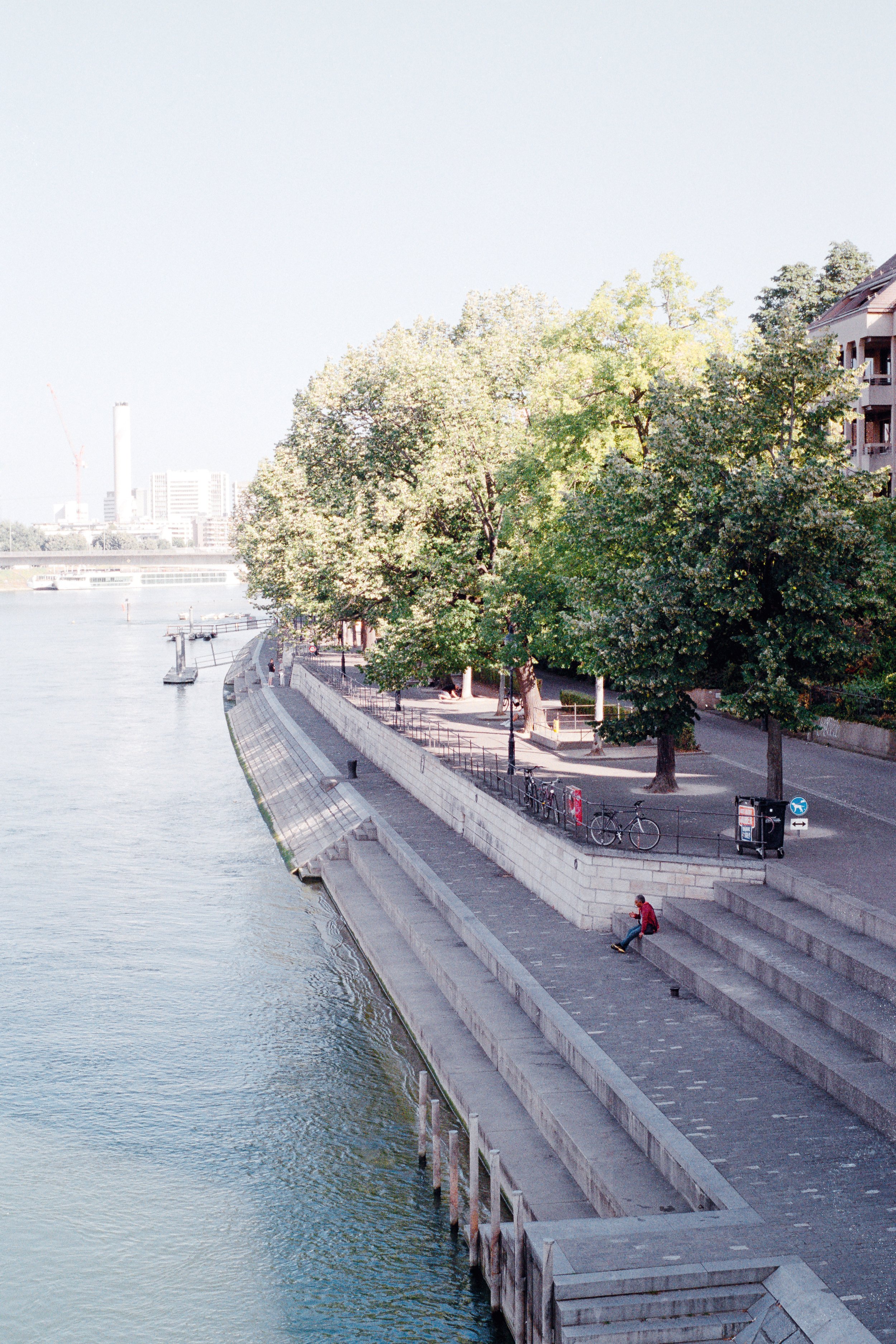 A promenade along the Rhine in Basel with large leafy trees, a person sitting on steps, and bicycles parked along the railing. Buildings and a river are visible in the background.