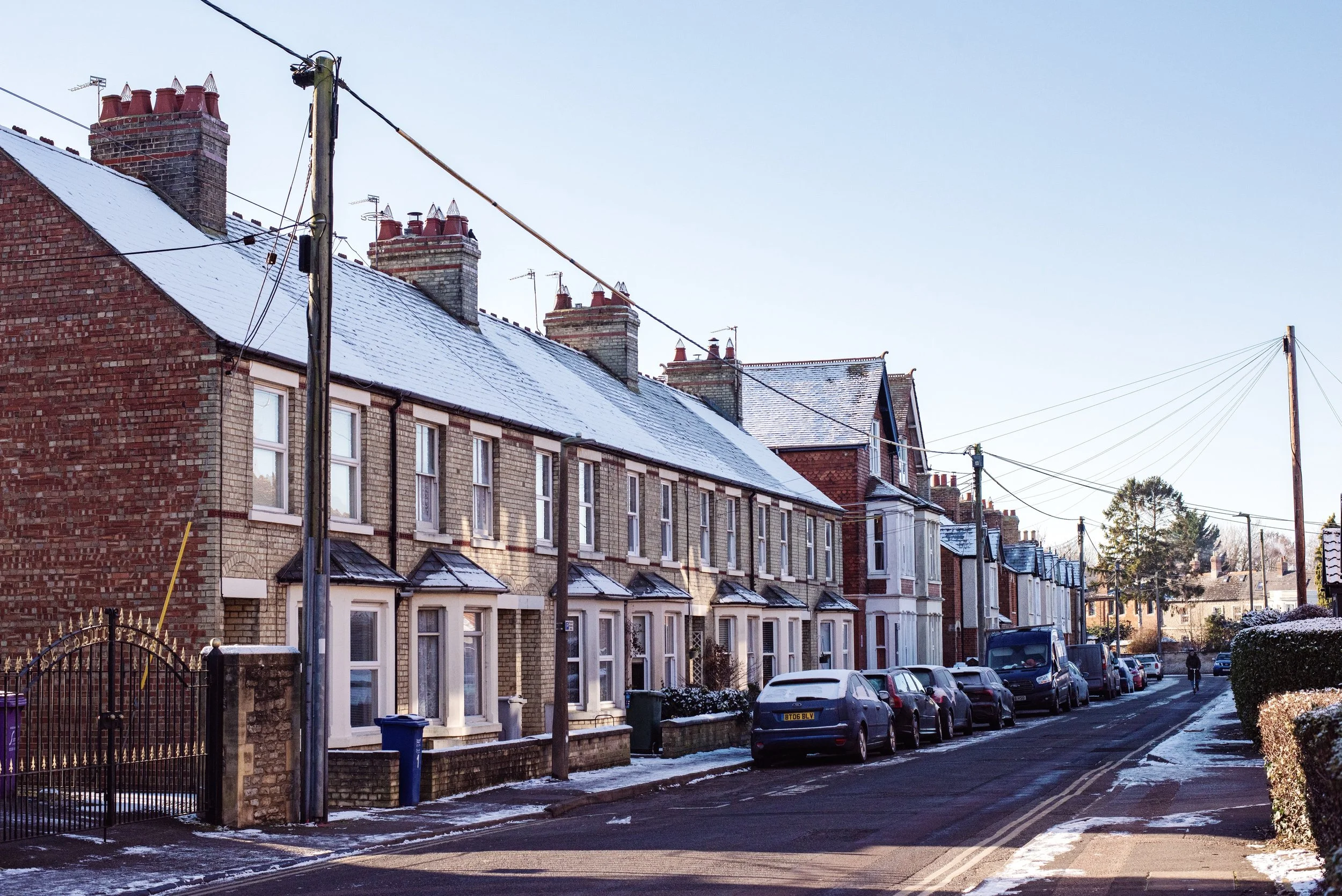 Residential street with terraced houses, some with snow on roofs, parked cars, and a person walking in the distance in Bicester, UK.