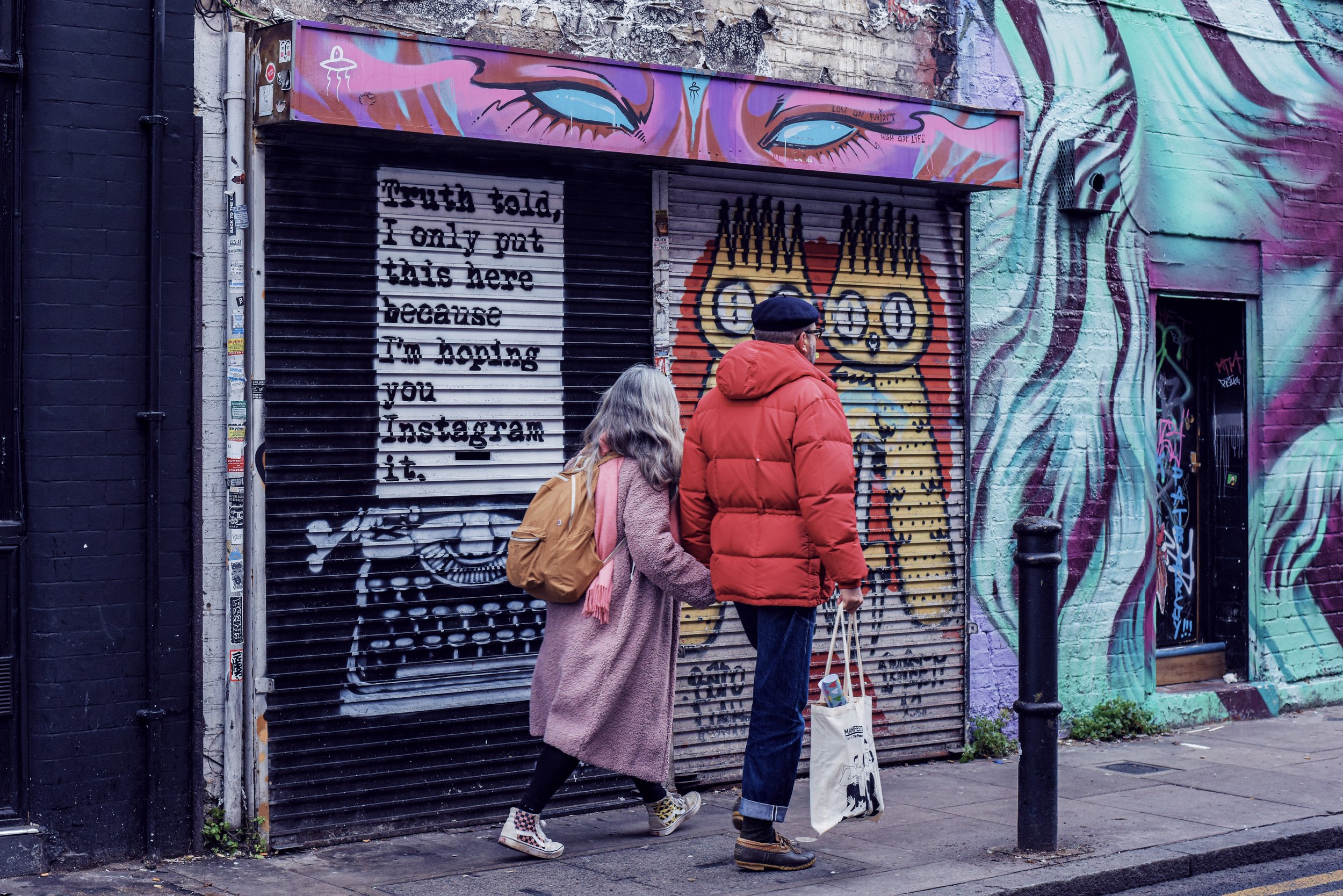 Two people walk past a mural on Brick Lane in London, with a roller shutter featuring a quote and graffiti art, including a colorful owl, an abstract face, and other street art elements.
