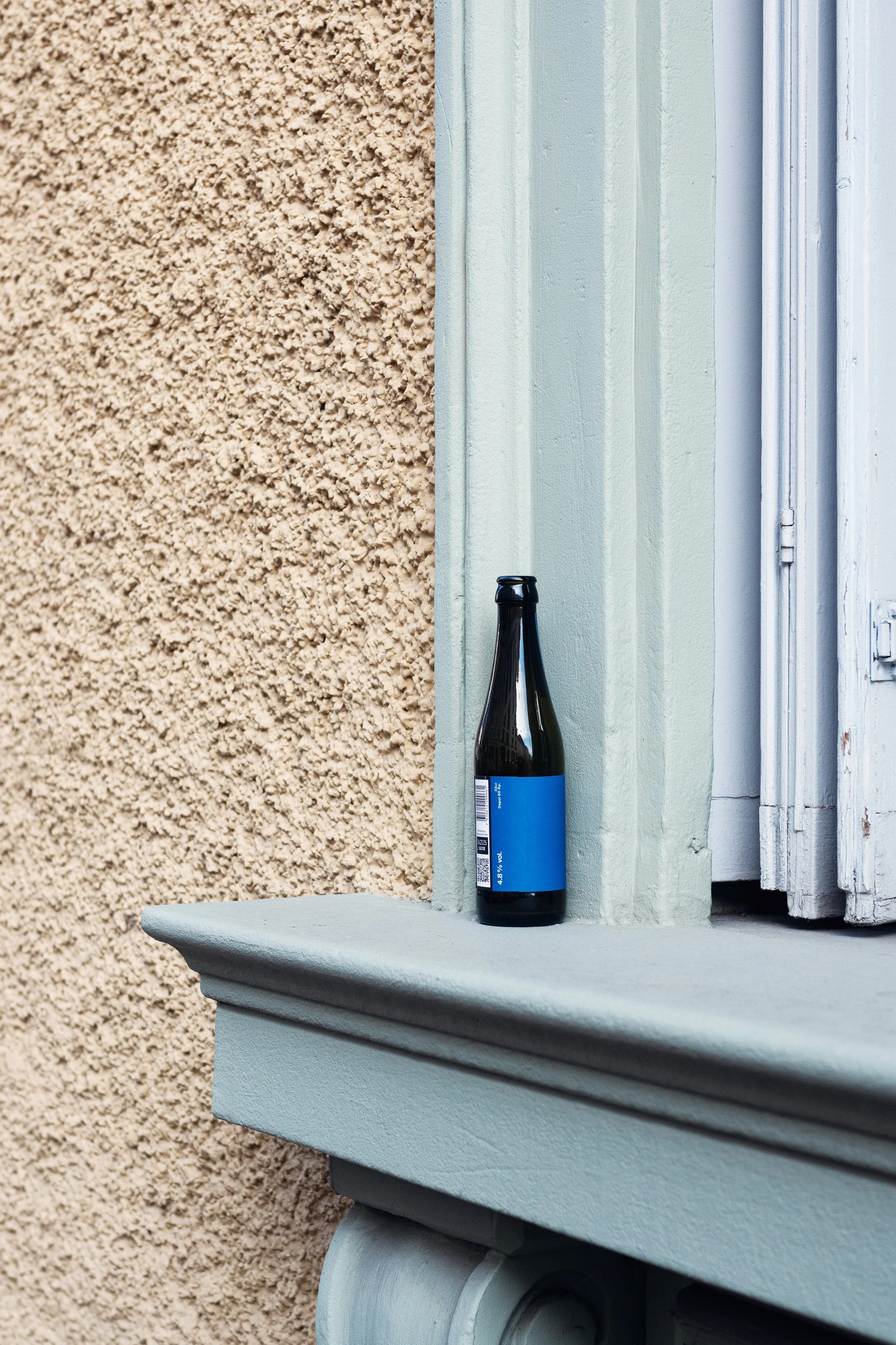 An empty dark glass bottle with a blue label placed on a window sill outside a building with beige and light green textured walls.