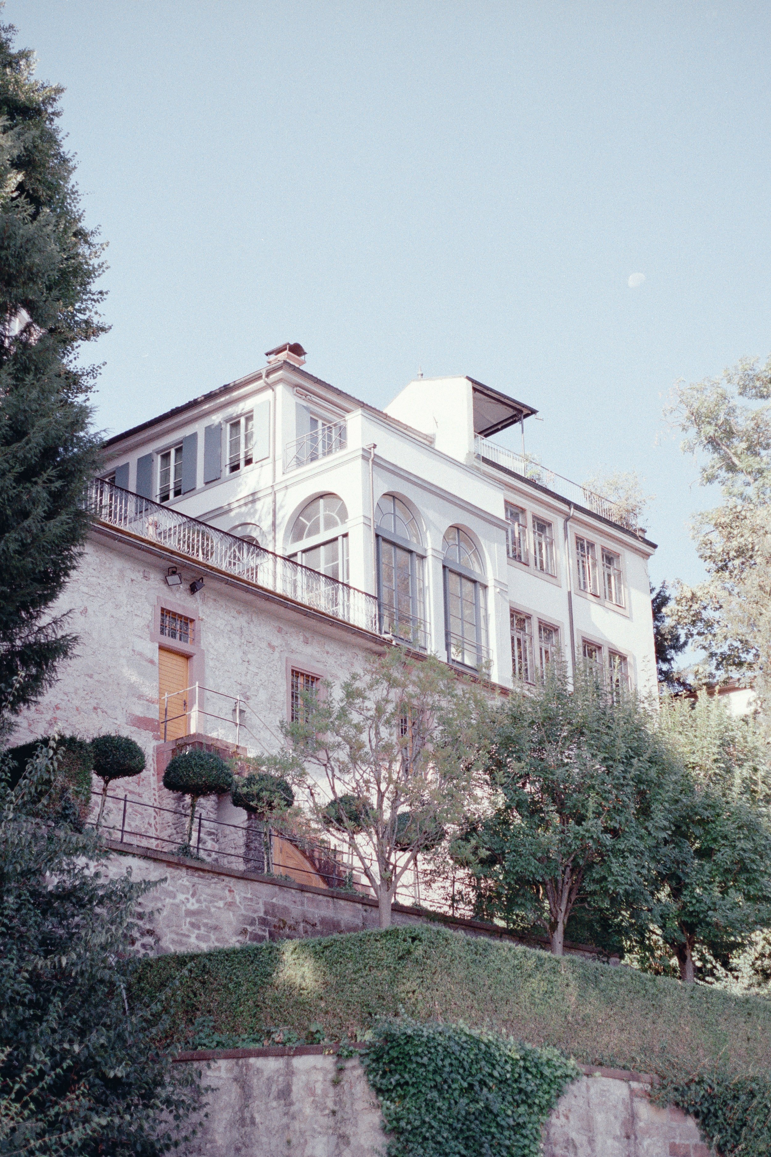 A multi-story house on a hillside surrounded by trees and shrubs, with a clear blue sky above and the moon visible in the sky in Basel, Switzerland.