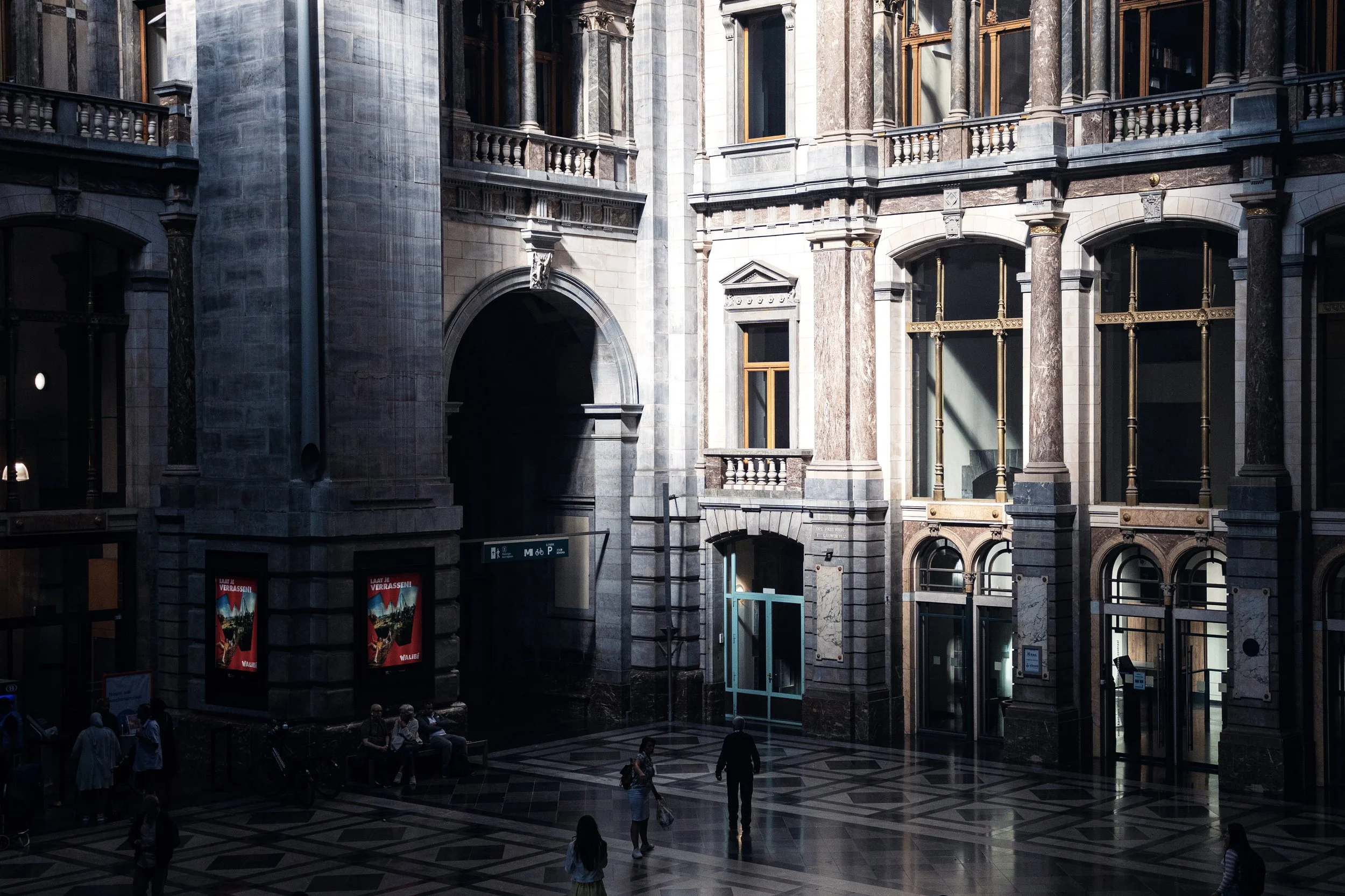 Interior view of the historic train station in Antwerp with tall arched windows, marble columns, and a patterned tiled floor, with a few people sitting and walking.