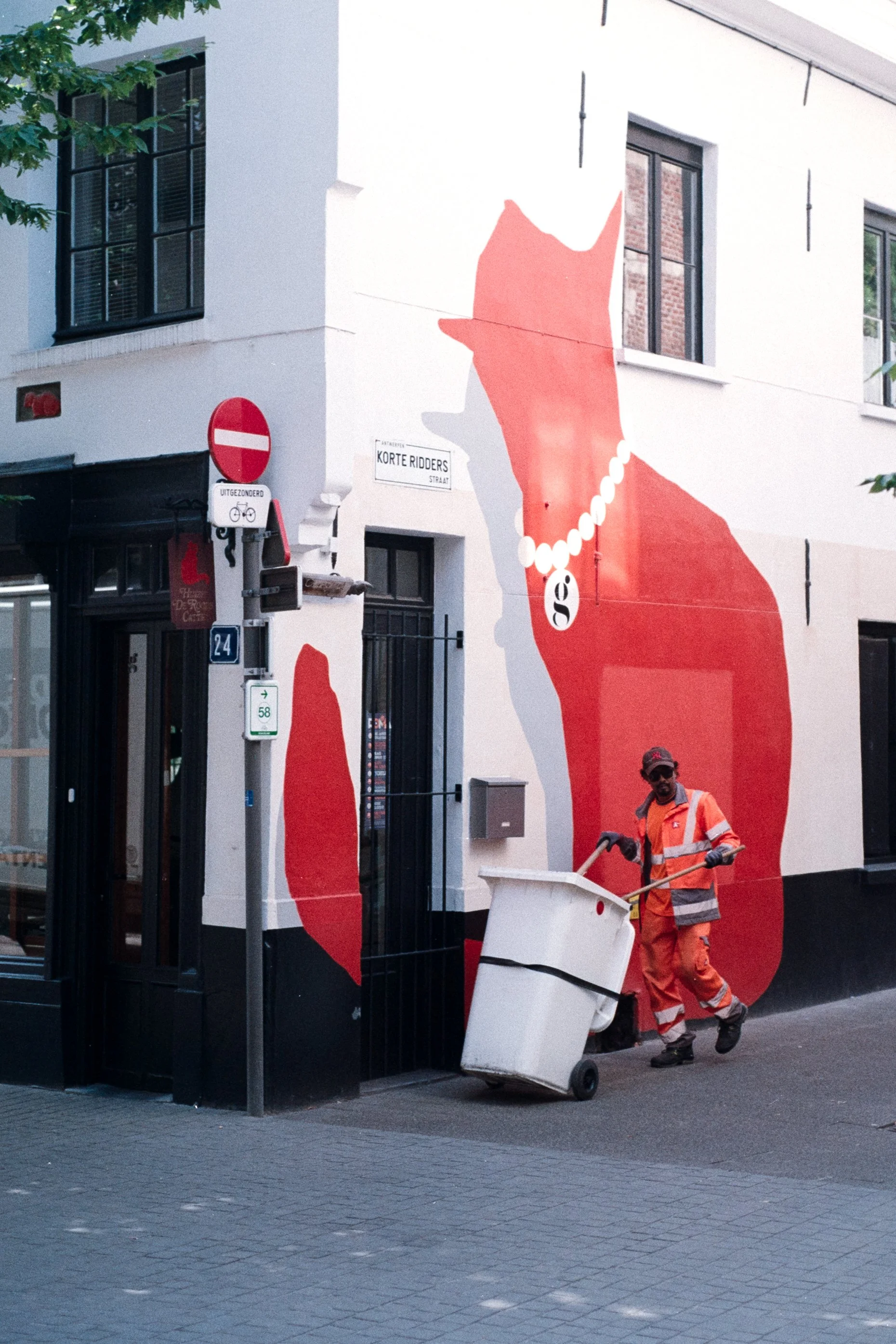 A man in orange reflective work clothing pushing a white trash bin on a city sidewalk in Antwerp, Belgium. The building behind him has a large mural of a red cat wearing a pearl necklace painted on the wall.