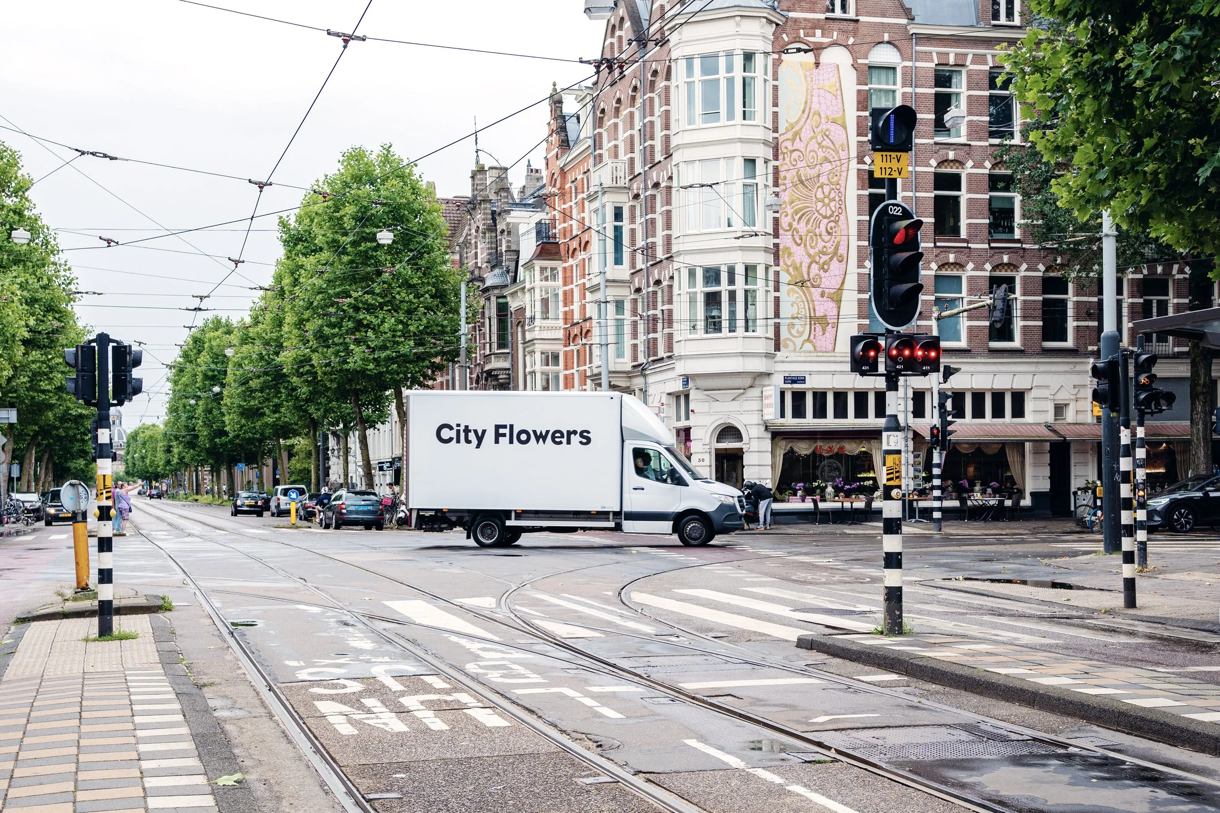 City street scene in Amsterdam, The Netherlands, with a moving white delivery truck labeled "City Flowers," tram tracks, traffic lights, a flower shop with outdoor seating in a multi-story brick building, and green trees lining the sidewalk.