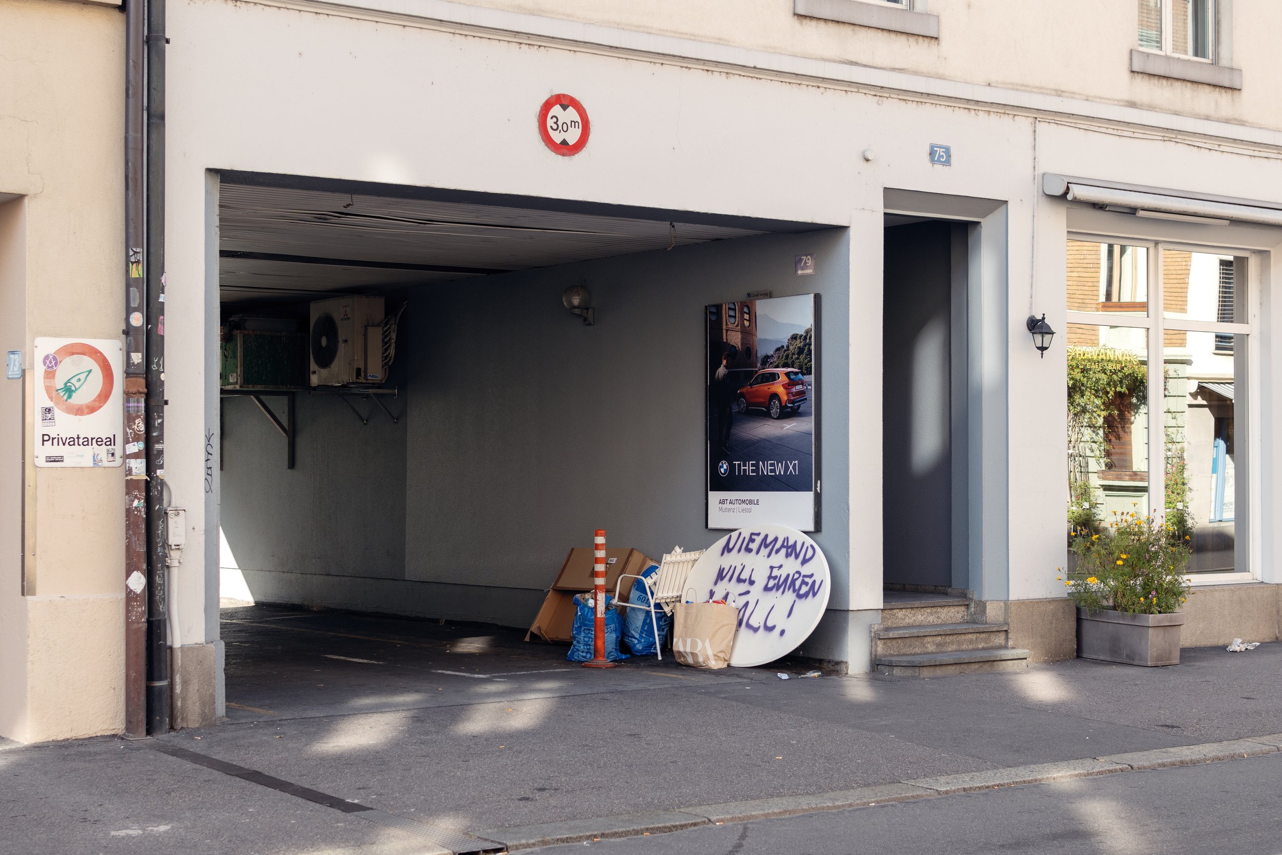 Side view of a building with a garage under a sign indicating a 3-meter height limit, containing boxed items, a traffic cone, a protest sign, and posters on the wall in Basel, Switzerland.