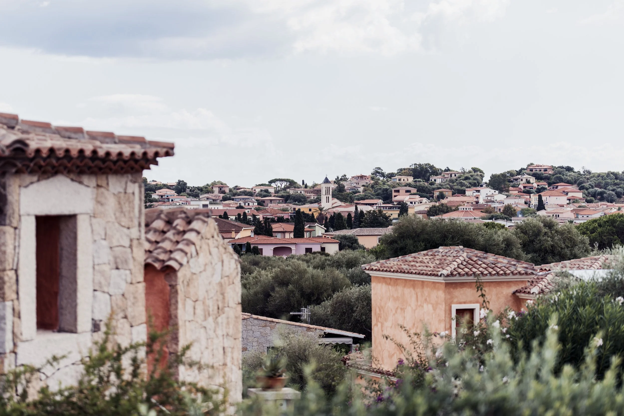 Scenic view of hillside houses with terracotta roofs, surrounded by greenery and trees, under a cloudy sky in San Teodoro, Sardinia.