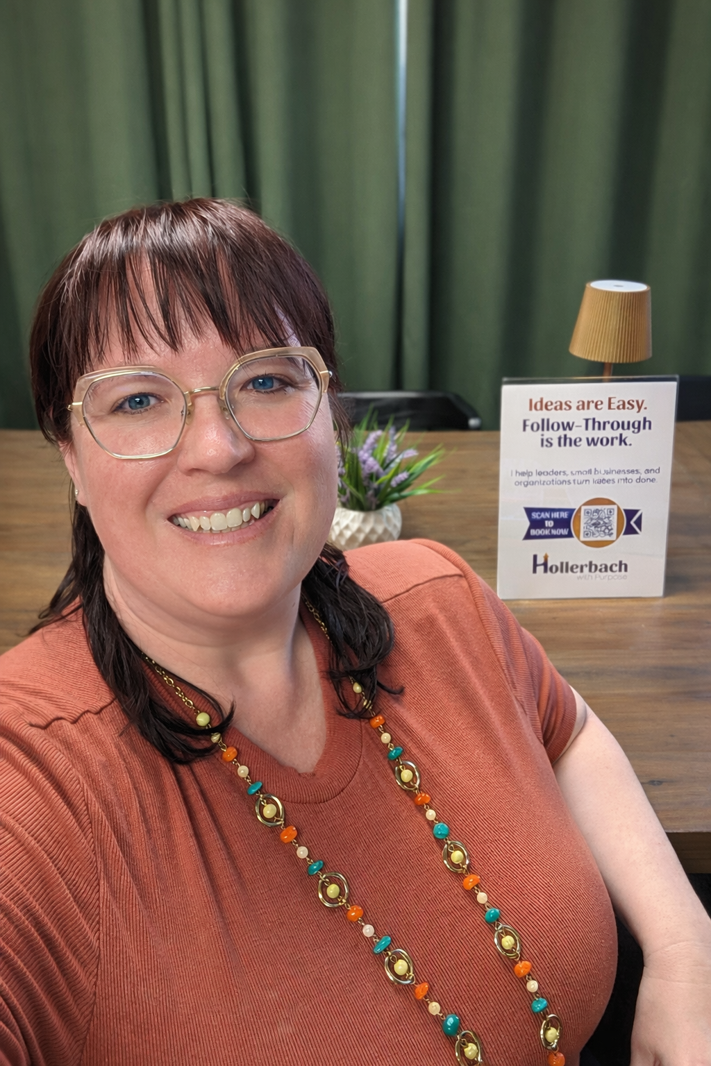 A woman with glasses and dark hair smiling at the camera, sitting at a wooden table with a sign, purple flowers, and a small lamp in the background.