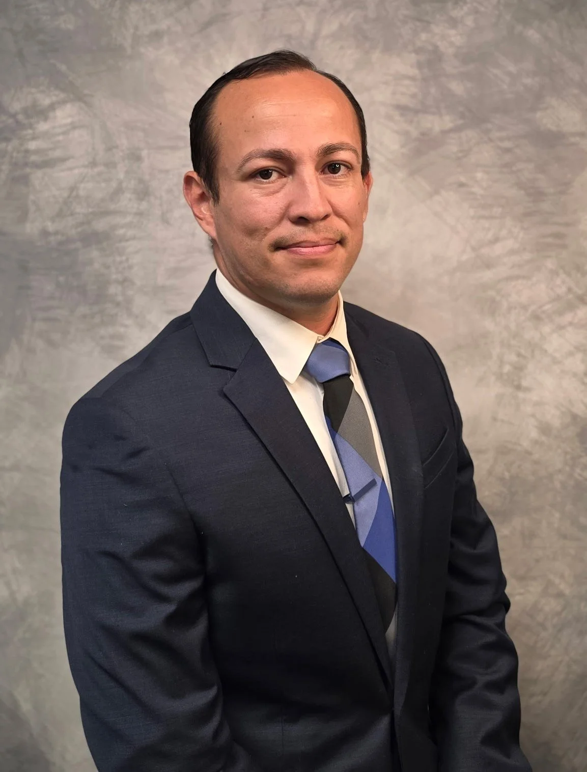 A man in a dark suit and a white shirt with a multi-colored striped tie, posing against a gray textured background.