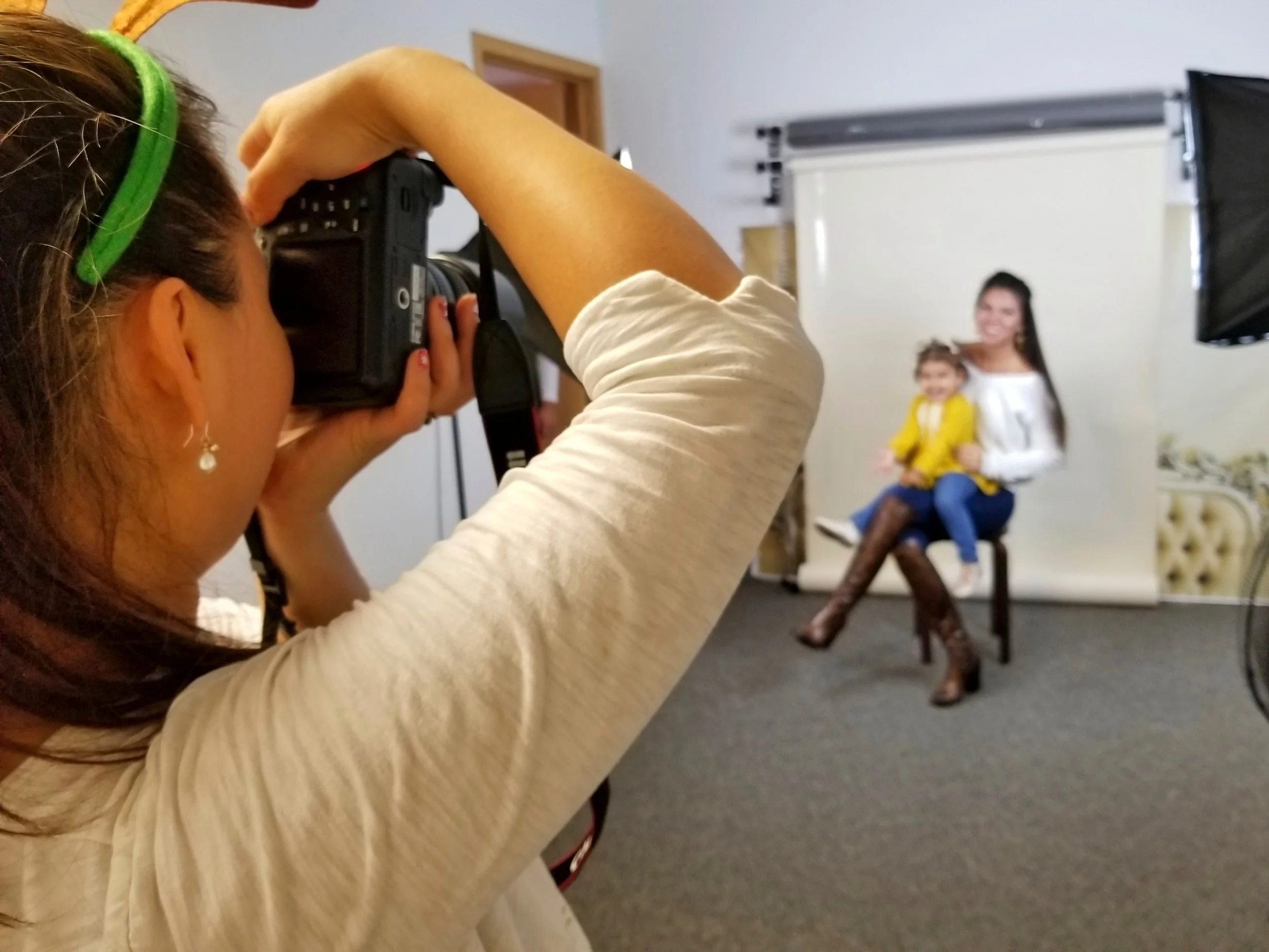 A woman in a white shirt and black pants is sitting on a chair with a small girl on her lap, in front of a white backdrop, being photographed by a photographer taking a picture with a Canon camera.