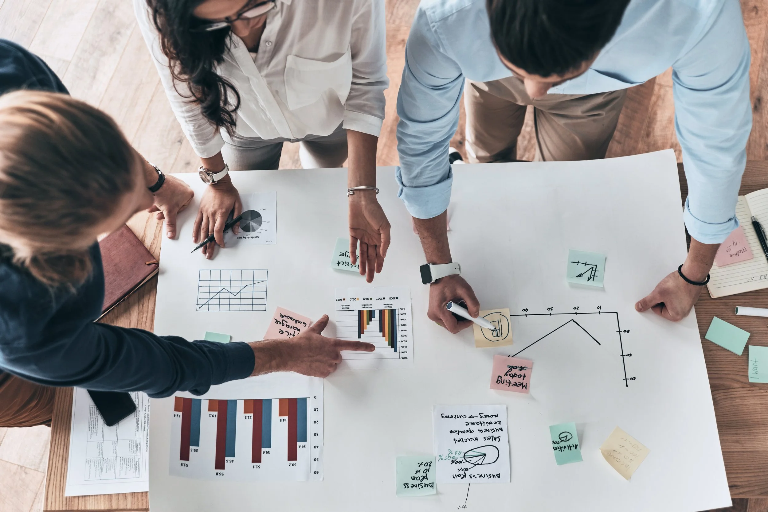 Group of four people working together on a business project at a table with charts, graphs, sticky notes, and documents.