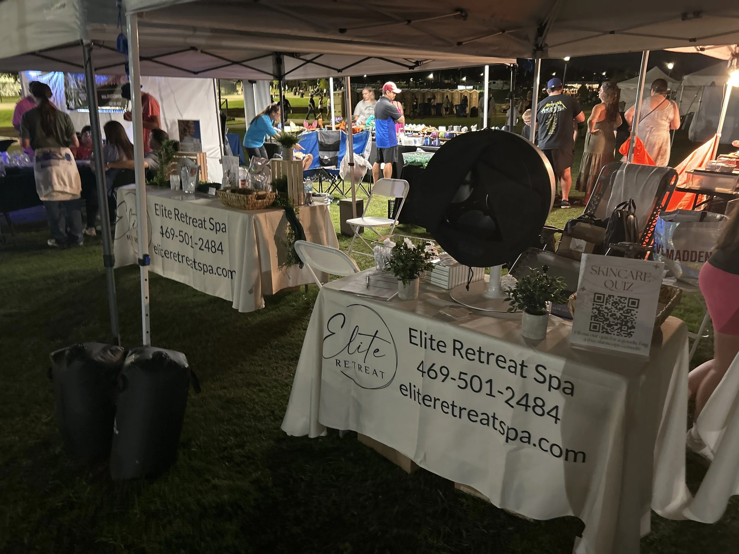 Nighttime outdoor event with vendor booths under white tents, featuring a spa booth with promotional banners, tables, chairs, and people browsing and chatting.