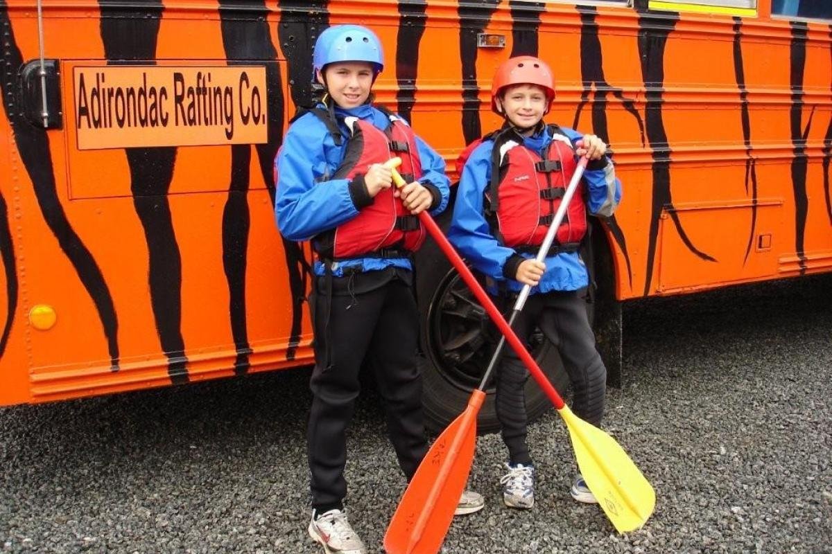 Two children dressed in outdoor gear and helmets holding paddles, standing in front of an orange striped vehicle with a sign that reads 'Adirondac Rafting Co.'