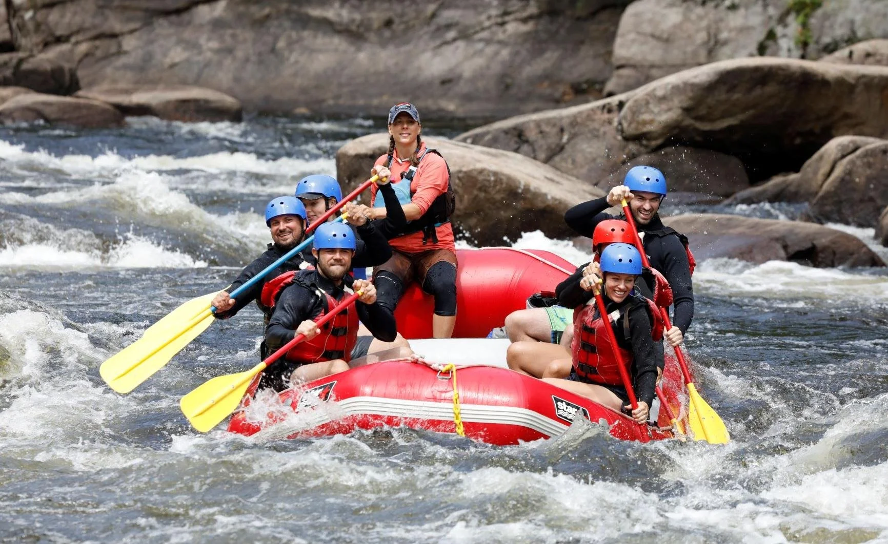 A group of six people white water rafting on the Hudson River, wearing helmets and life jackets, paddling together, with rocky surroundings.