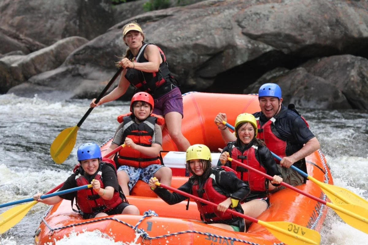 A group of six people, including children and an adult, are whitewater rafting on a river. They are wearing life jackets and helmets, and some are holding paddles while smiling and having fun.