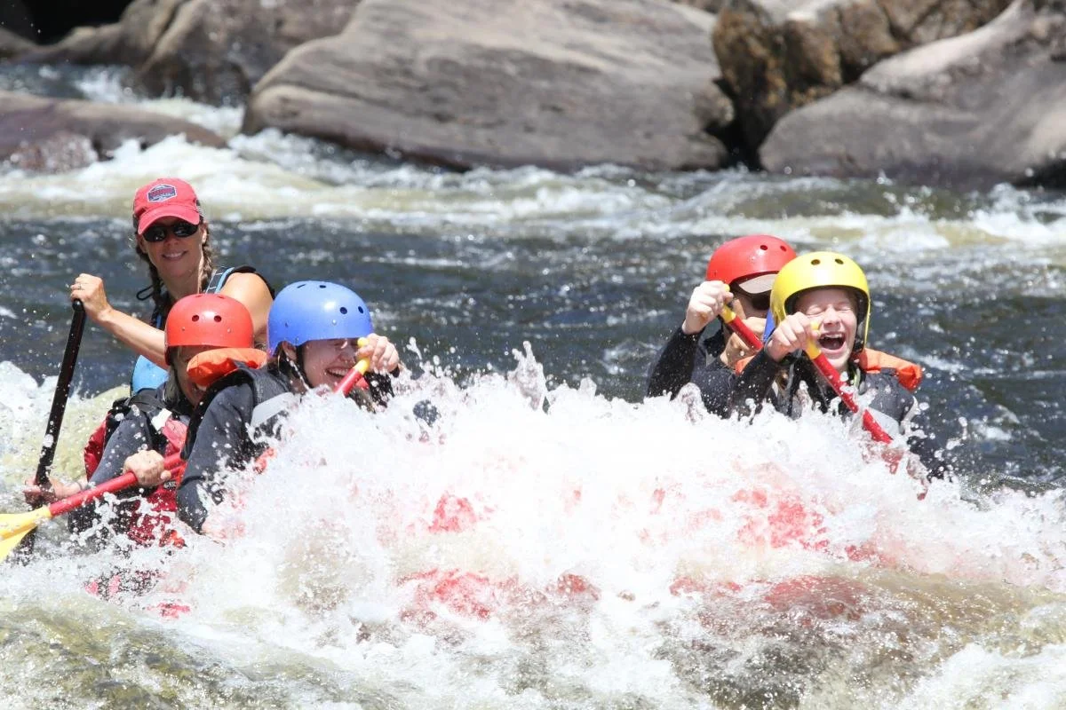 A group of five people white-water rafting in the Hudson River, wearing colorful helmets and life jackets, with rocks in the background.