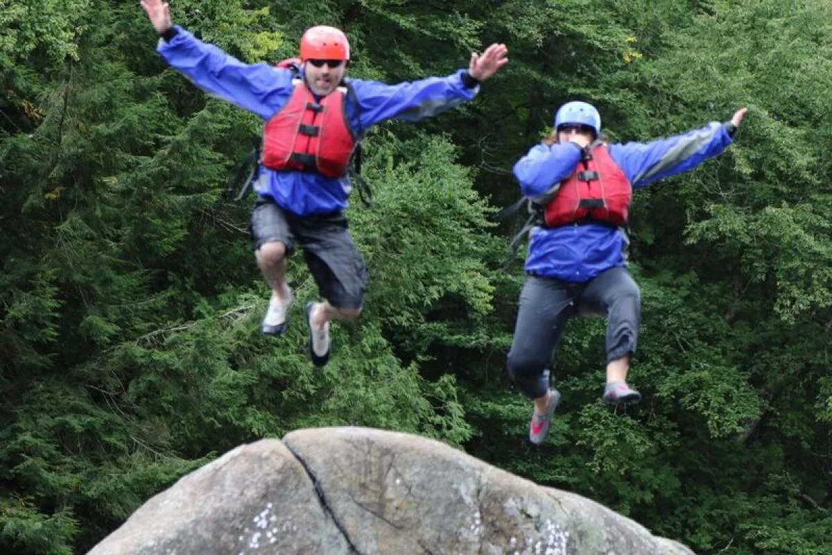 Two people in blue jackets, red life vests, and helmets jumping off a rock into a river in a forested area.