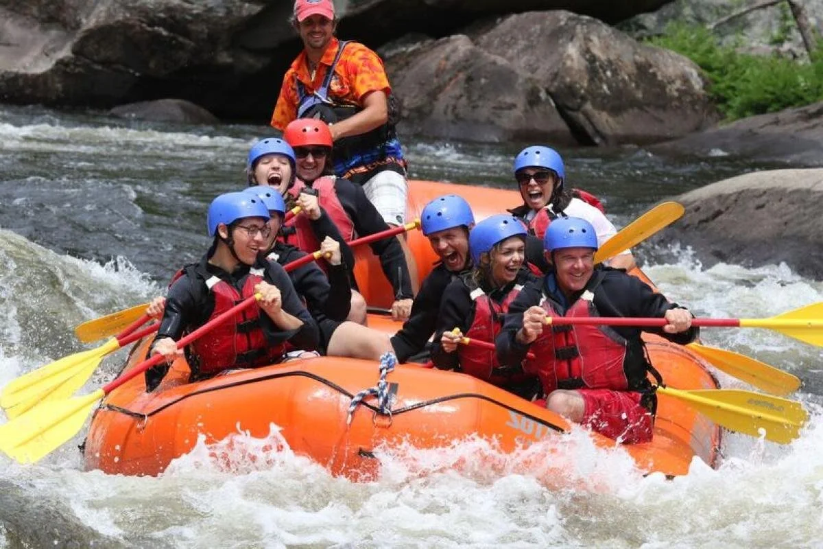 A group of people white water rafting in an orange raft, wearing safety helmets and life jackets, and smiling while paddling through rapids.