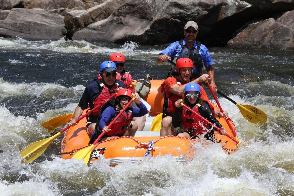 Group of six people white water rafting on a river surrounded by rocks, wearing life jackets and helmets, with one person standing at the back and the others paddling.