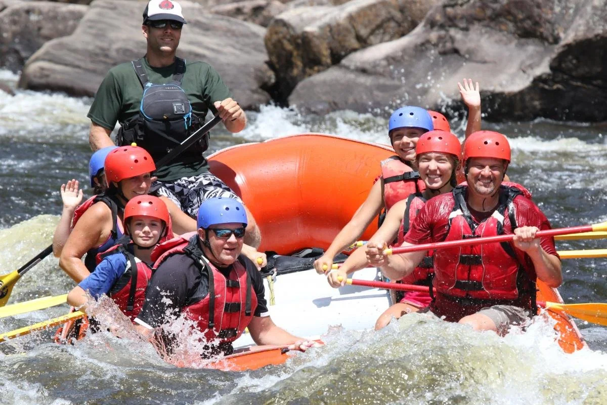 Group of six people and one guide white water rafting on the Hudson River, all wearing life vests and helmets, with rocks in the background.