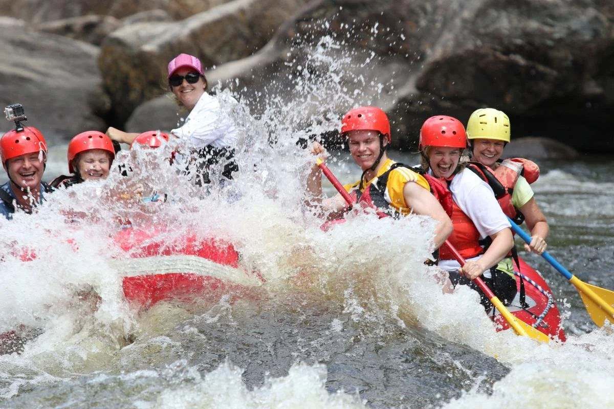 Group of people white water rafting, wearing helmets and life jackets, smiling and splashing in river with rocks in background.