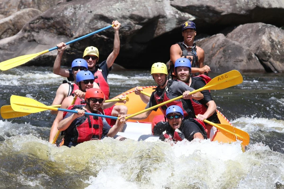 A group of six people white water rafting, wearing helmets and life jackets, with smiling and excited expressions, paddling through a river with rocks and rapids.