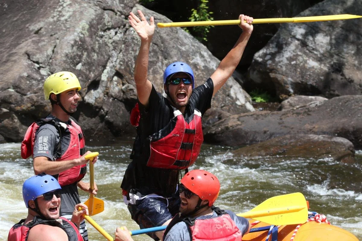 Four men in life jackets and helmets white water rafting on a river, with one man raising his arms and shouting in excitement.