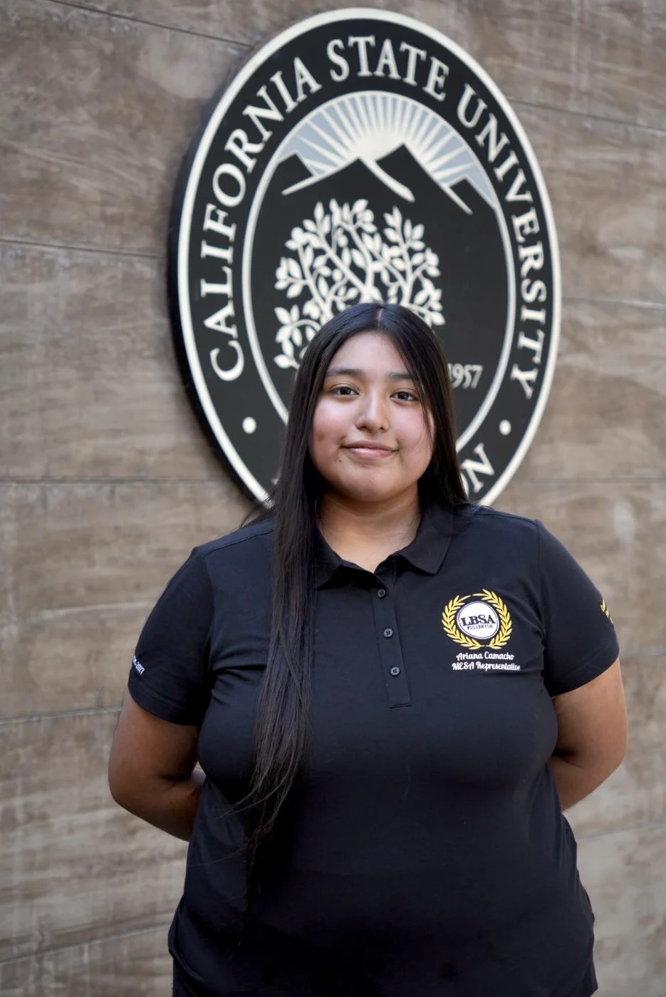 A young woman with long brown hair and a black polo shirt, smiling, standing in front of a large circular logo that reads 'California State University, Fullerton.'