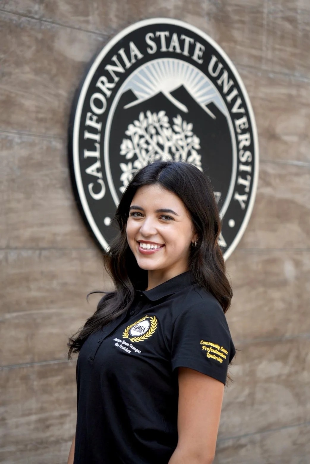 A young woman smiling in front of the Cal State Fullerton logo, wearing a black polo with her name and titles embroidered on it.