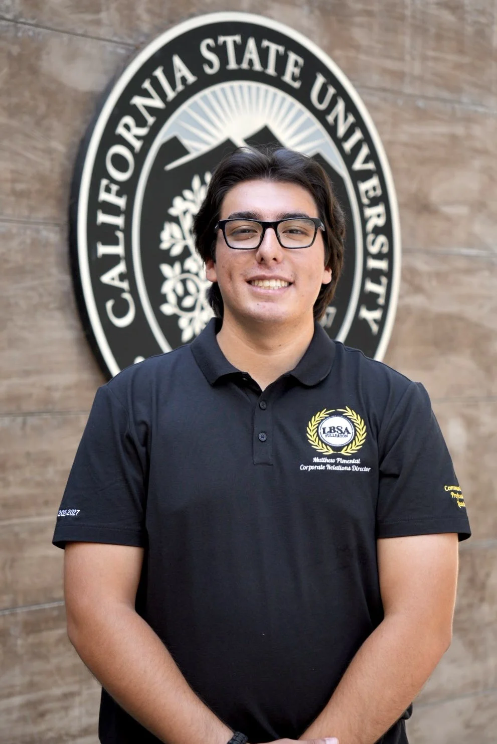 Young man with glasses and dark hair standing in front of a California State University sign, wearing a black polo shirt with embroidered text.