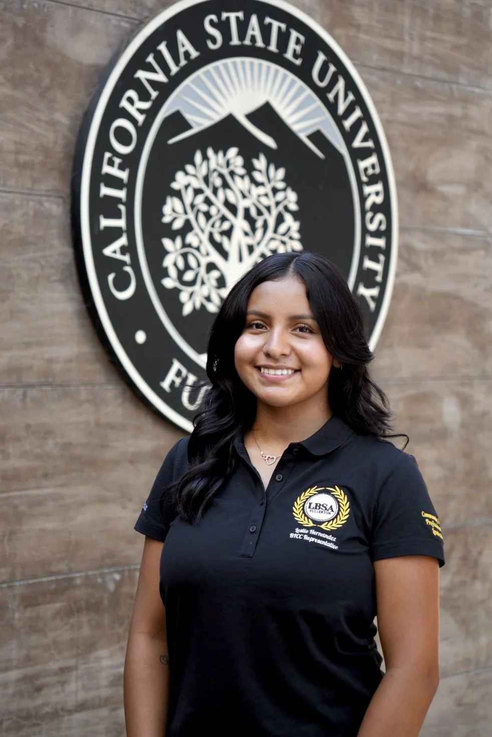 A young woman with long dark hair and a beige necklace, smiling in front of a California State University sign.