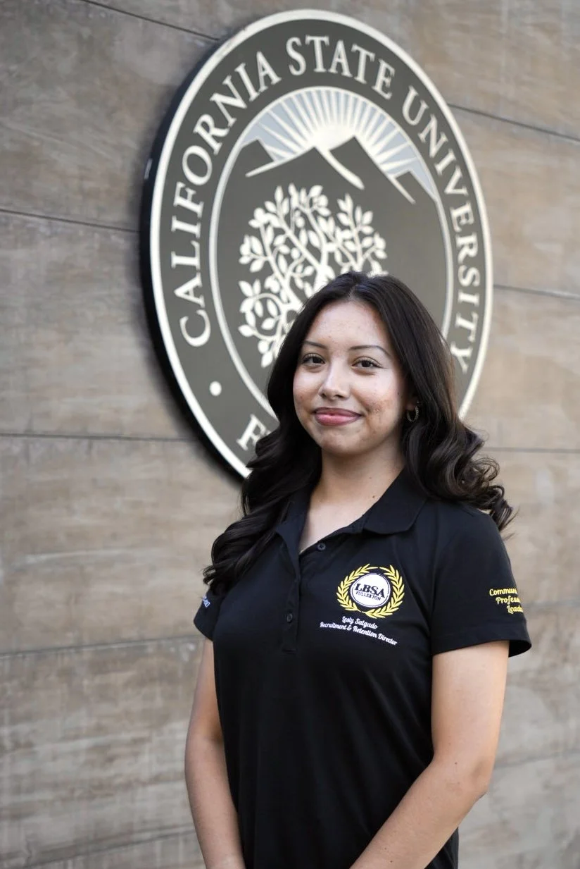 A young woman with long wavy red hair smiling in front of a large black and white California State University logo. She is wearing a black polo shirt with embroidered text and a crest.