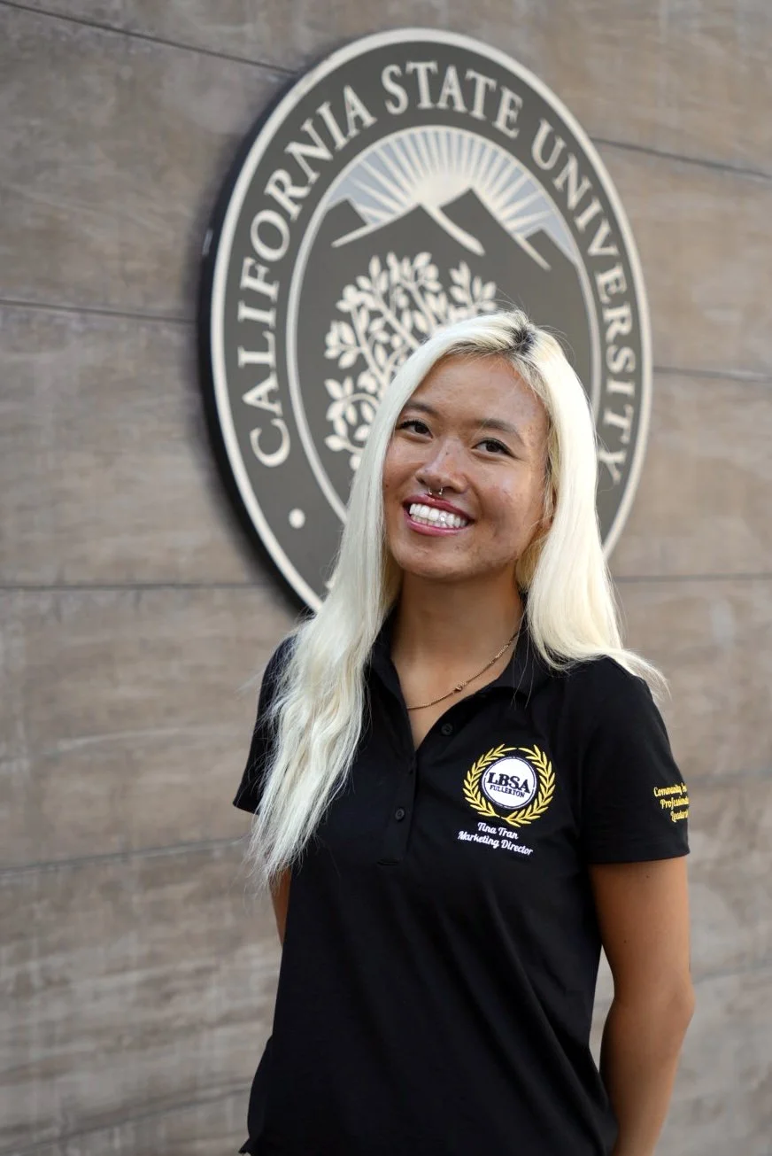 A smiling woman with long brown hair and makeup, wearing a black shirt with embroidered text and a logo, standing in front of a wooden wall and a California State University sign.