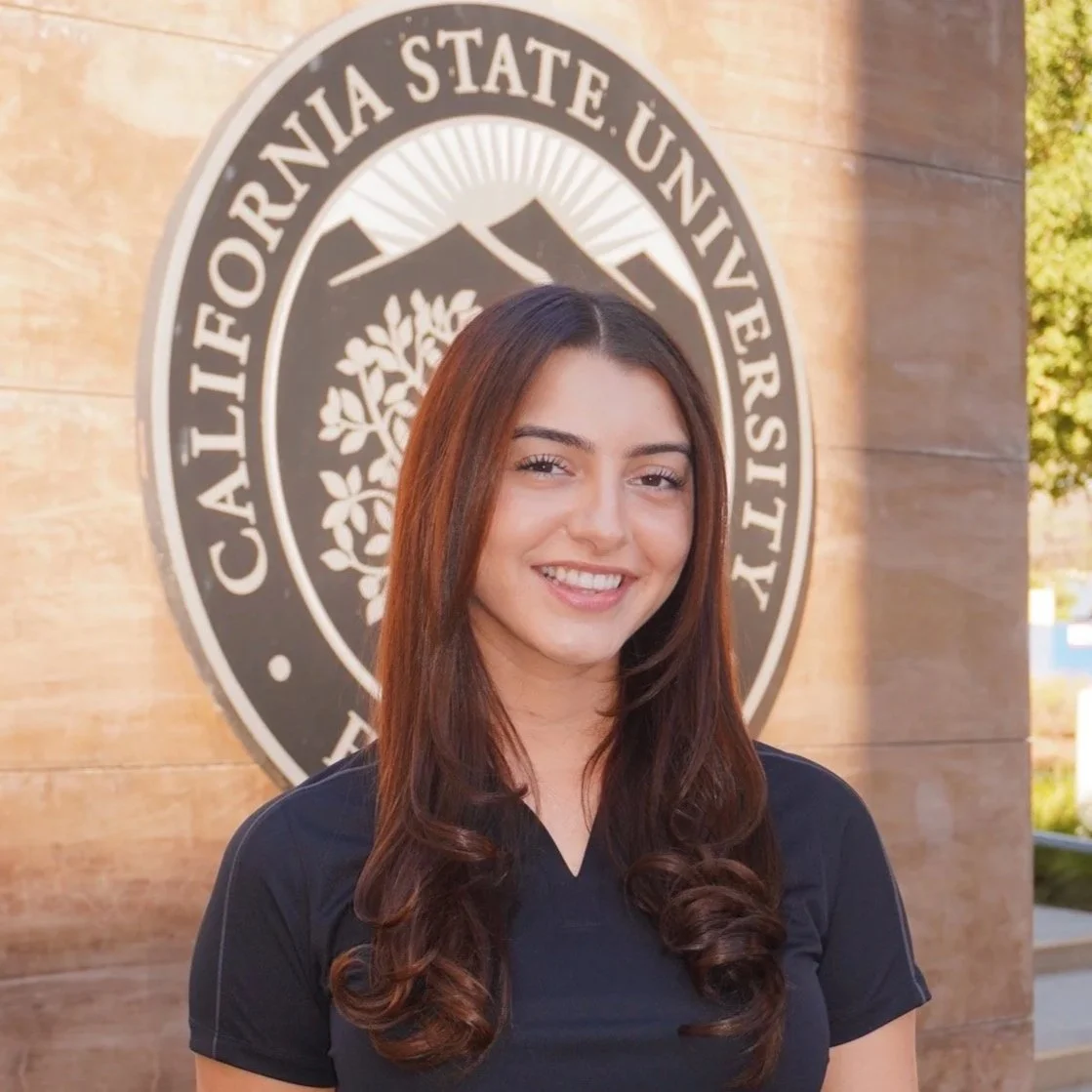 Young woman smiling, wearing a black polo shirt, standing in front of a circular California State University, Fullerton emblem on a wood-paneled wall.