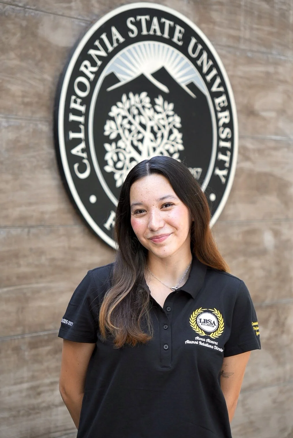 Young woman smiling, wearing a black polo shirt, standing in front of a circular California State University, Fullerton emblem on a wood-paneled wall.