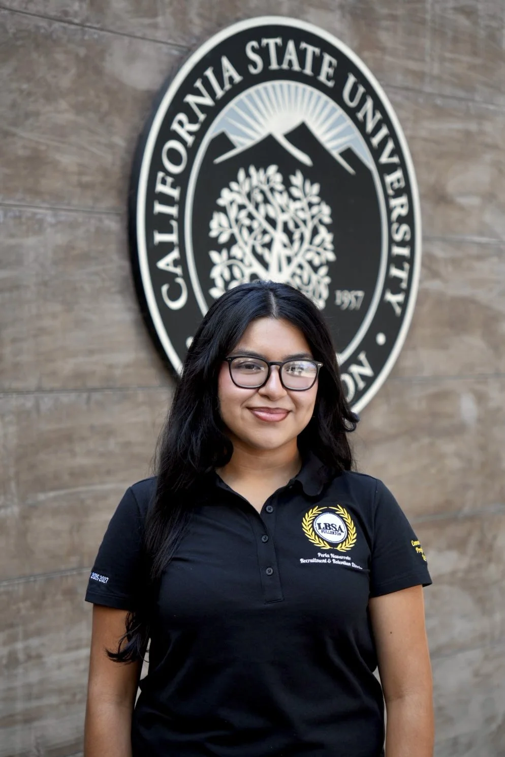 A young woman smiling in front of a California State University sign, wearing a navy blue polo shirt with her name and title embroidered on it.