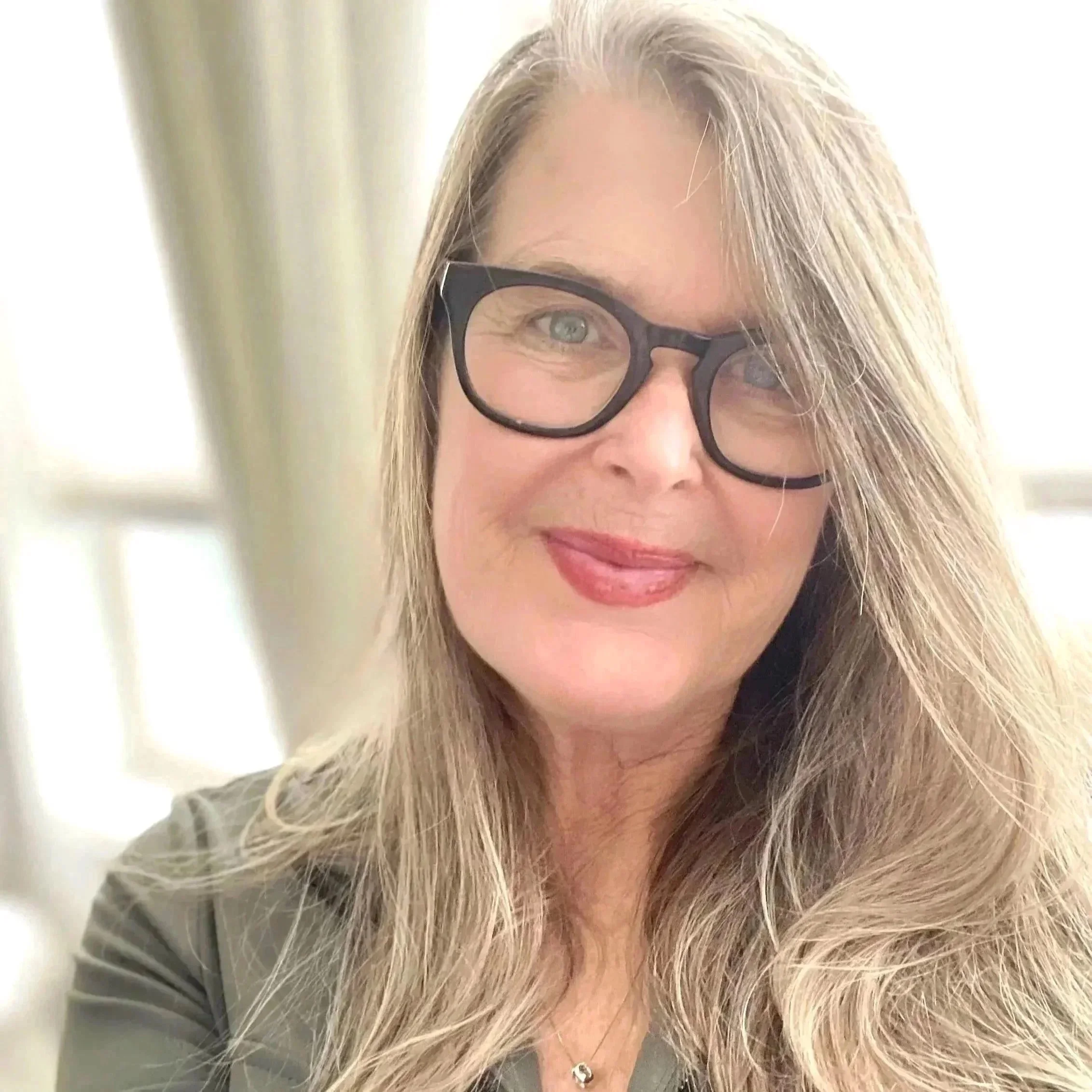 Close-up of a woman with long, blonde hair wearing black glasses, smiling slightly, with a blurred background of curtains and part of a gray shirt visible.