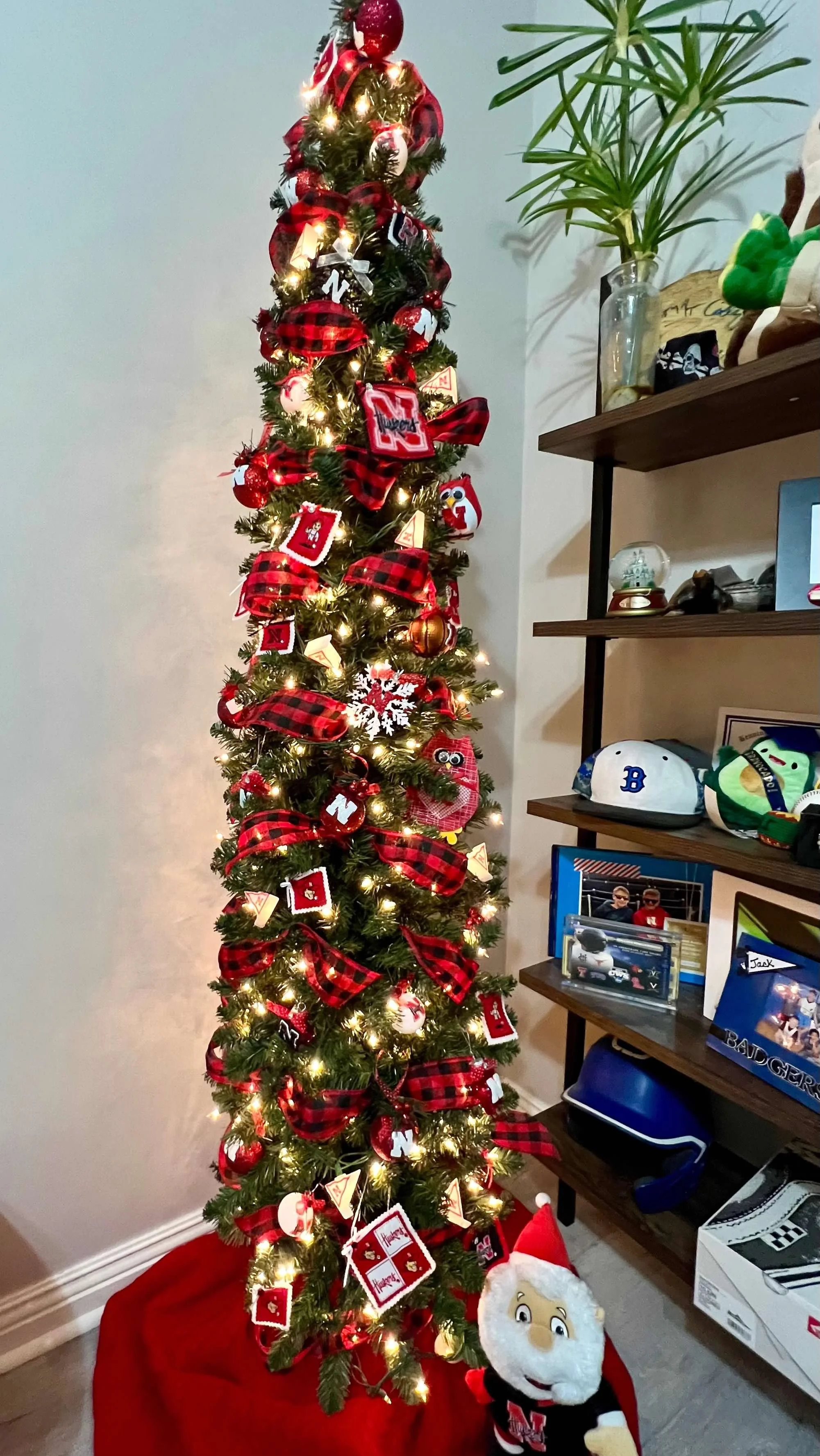 Tall Christmas tree decorated with red, black, and white ornaments, ribbons, and lights next to a shelf with memorabilia and a plush Santa Claus toy at the base.