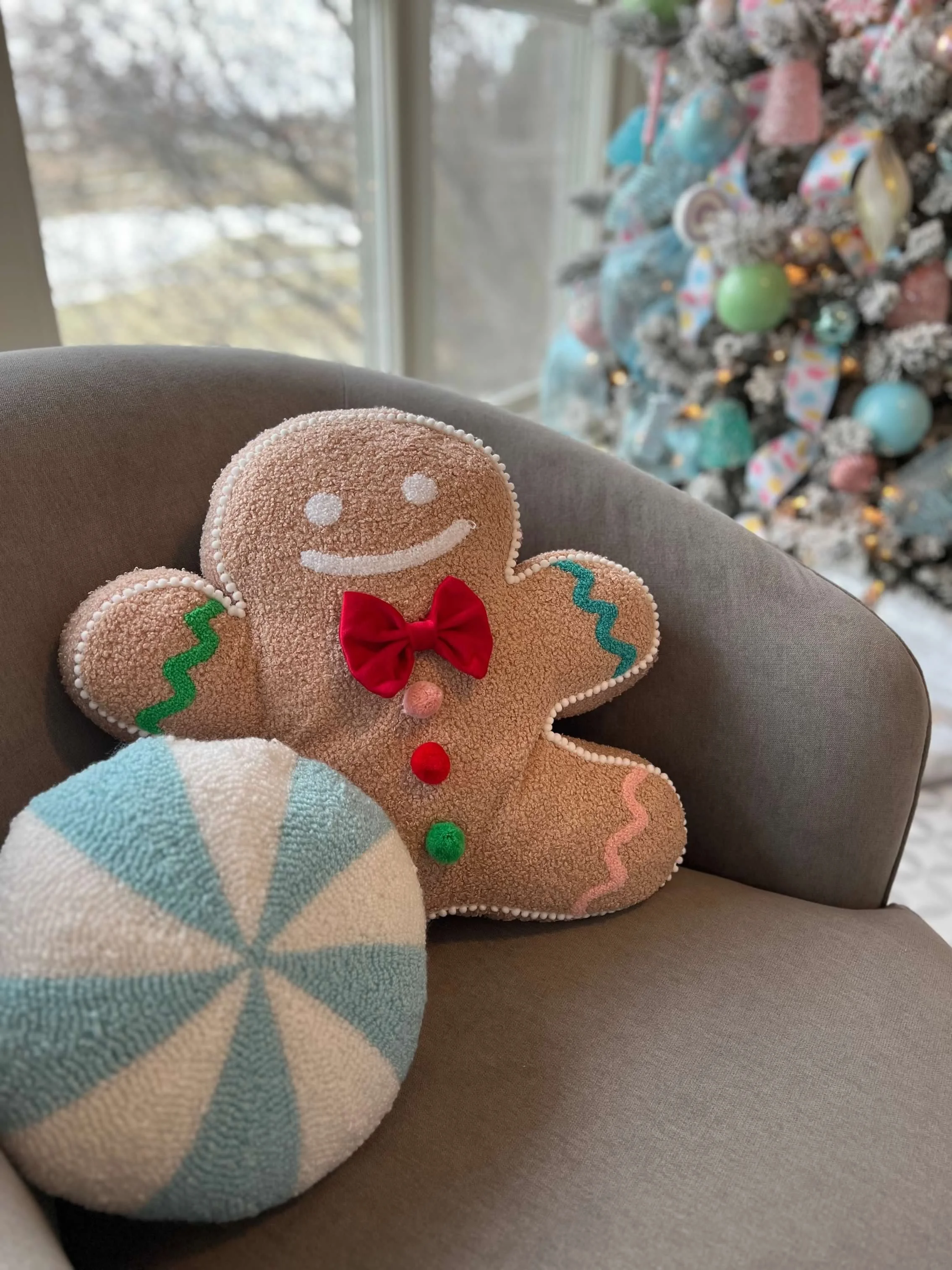 Decorative gingerbread man ornament and a round holiday pillow on a gray armchair with a decorated Christmas tree in the background.