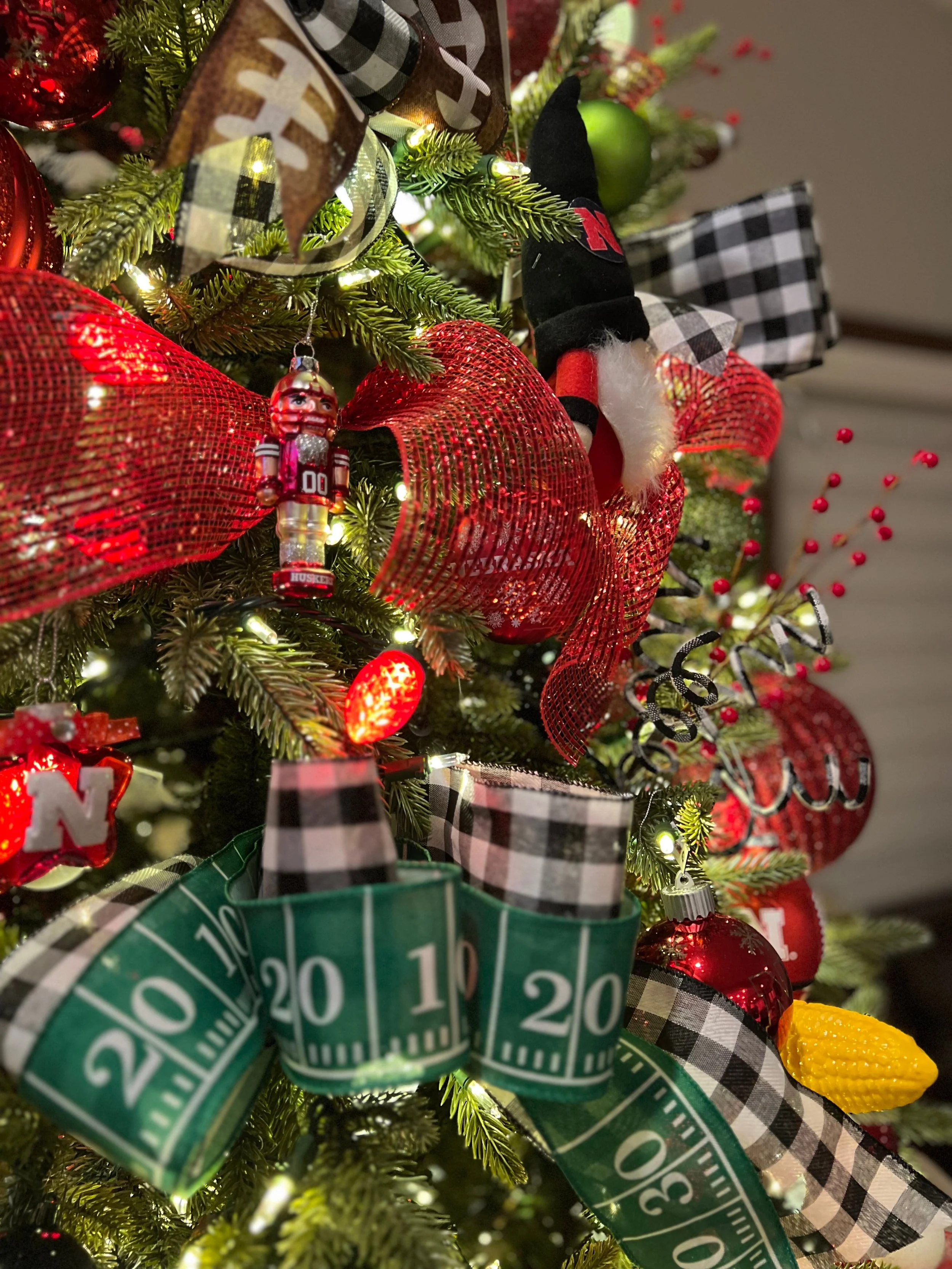 Close-up of a decorated Christmas tree with red and black plaid ribbons, a miniature football player ornament, a Nutcracker ornament, a yellow ornament, a green ornament, and a green measuring tape, with small white lights.
