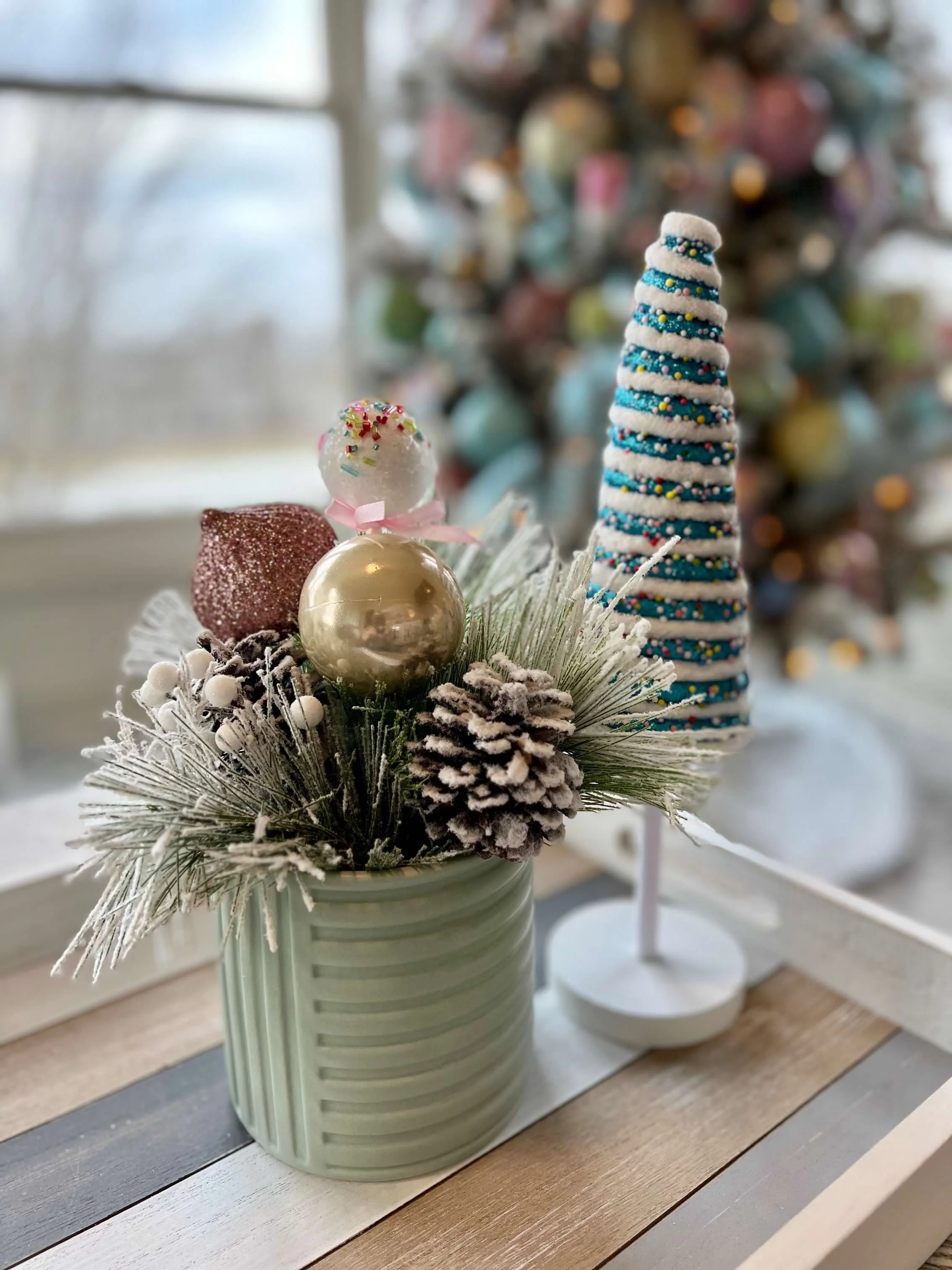 Christmas decorated centerpiece with pink, gold, and glittery ornaments, pine cones, artificial snow, and a white and blue tree cone, in a green vase on a wooden table near a window, with a decorated Christmas tree in the background.