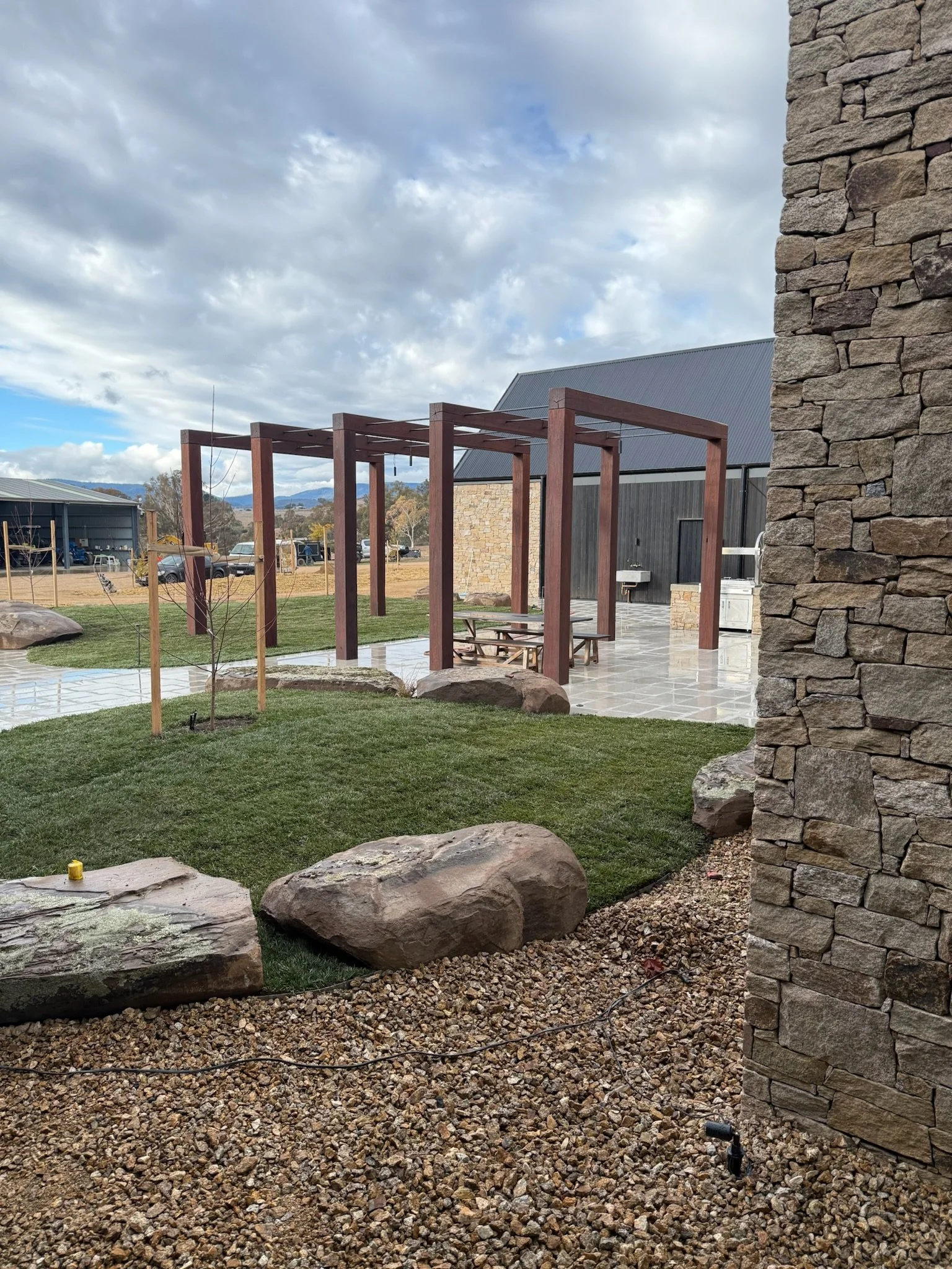 Outdoor patio area with wooden pergola, a picnic table, stone wall, grassy patch, large rocks, and gravel ground, under a partly cloudy sky.