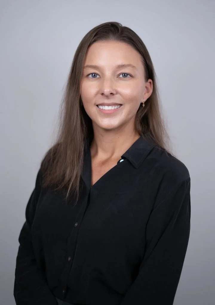 A portrait of a young woman with long brown hair, blue eyes, and a smile, wearing a black button-up shirt against a plain light gray background.