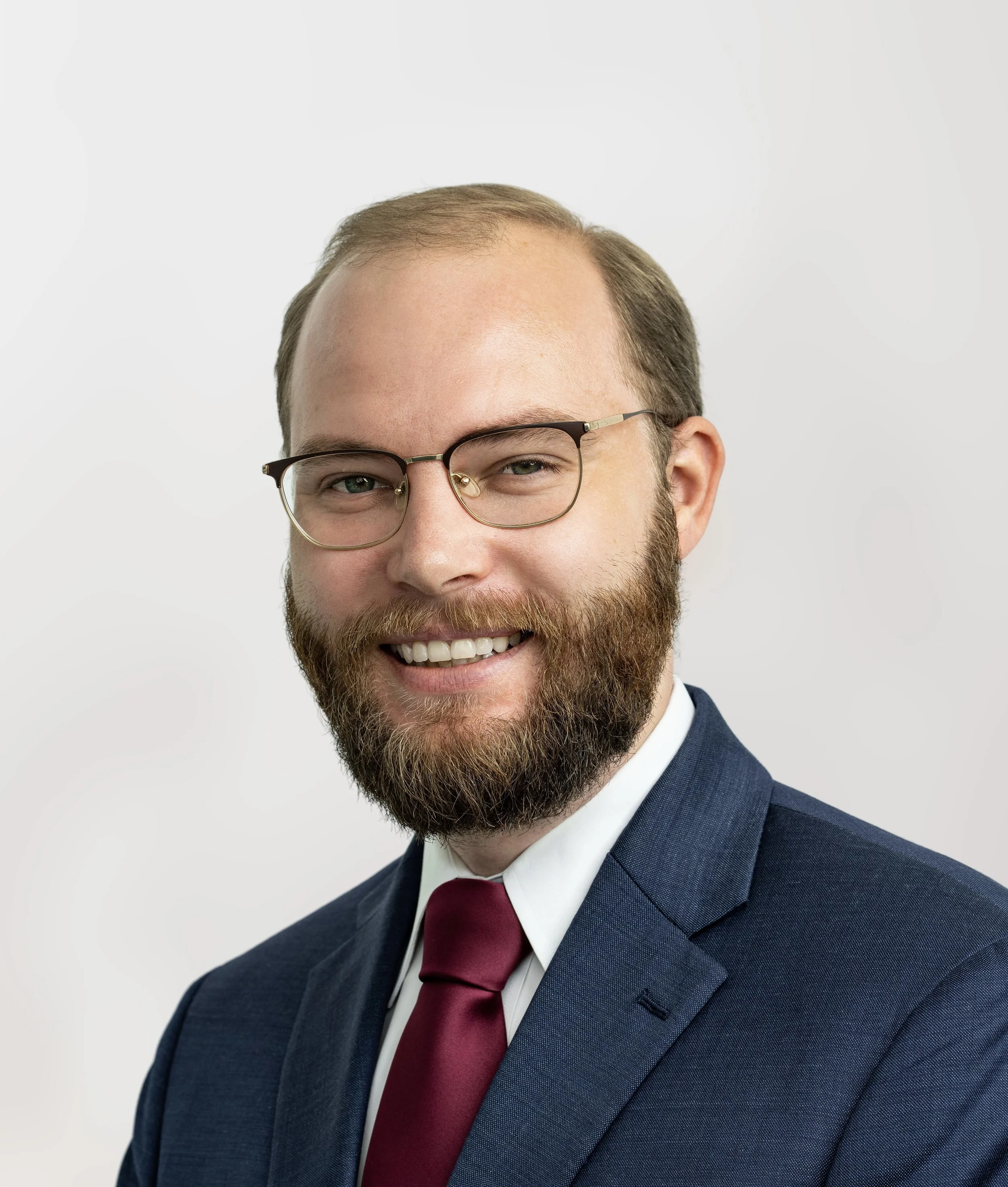 Portrait of a man with glasses, a beard, wearing a navy suit, white shirt, maroon tie, smiling against a plain white background.