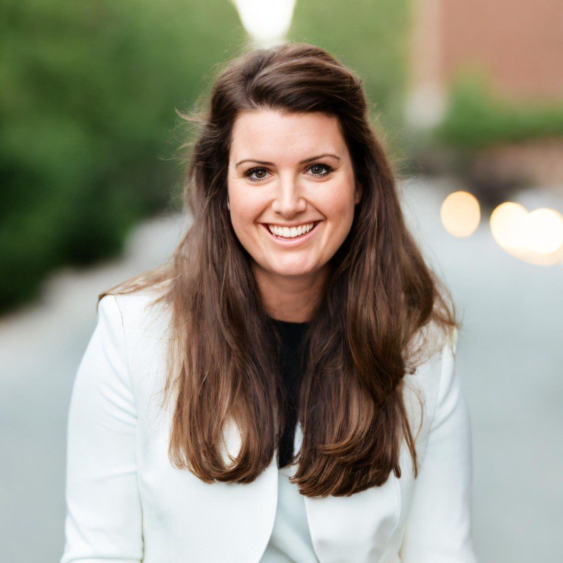A woman with long brown hair smiling outdoors, wearing a white jacket and black top.